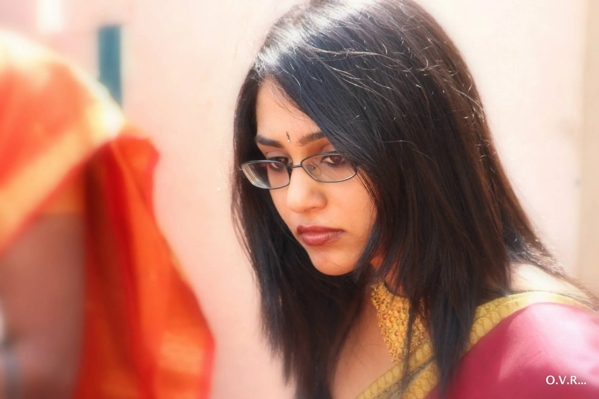 A woman with long black hair, glasses, and traditional Indian attire, looking downwards.
