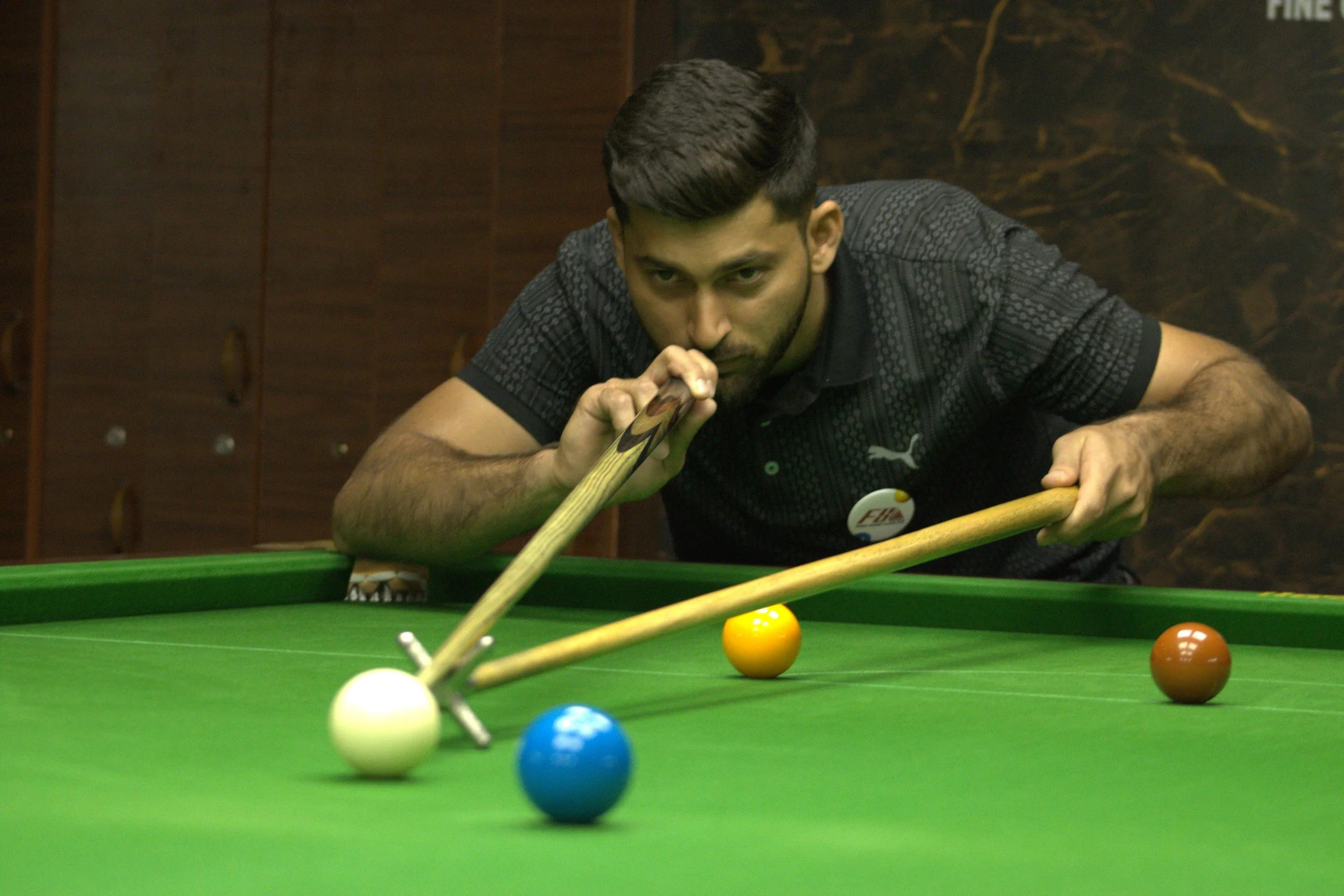 A man with dark hair and a beard playing snooker, aiming to hit the white cue ball with a cue stick, on a green snooker table. There are colored balls on the table, including yellow, blue, brown, orange, and yellow. The background features dark woode