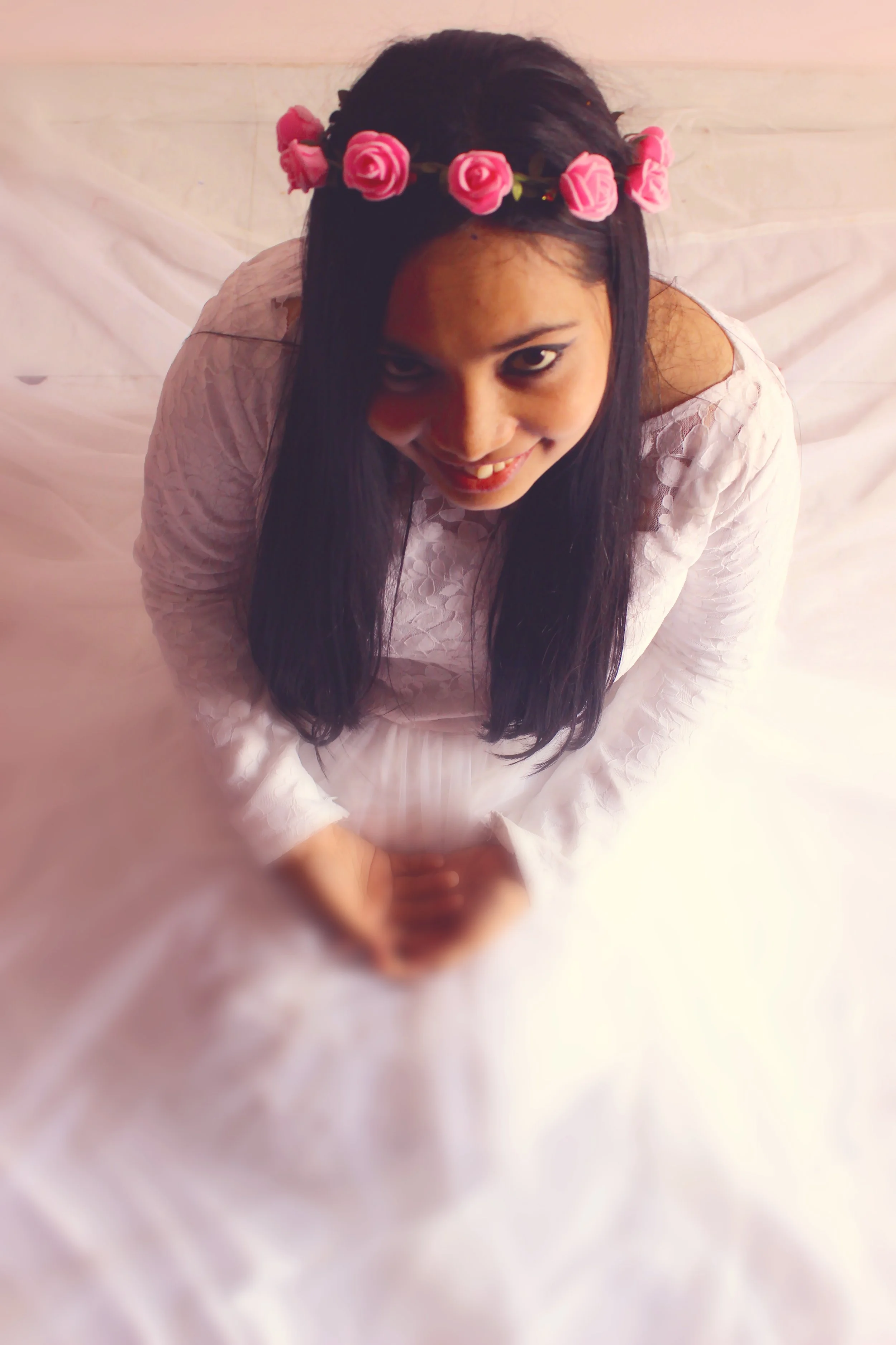 A woman with long dark hair wearing a white dress and a pink rose headband, looking up at the camera with a smile.