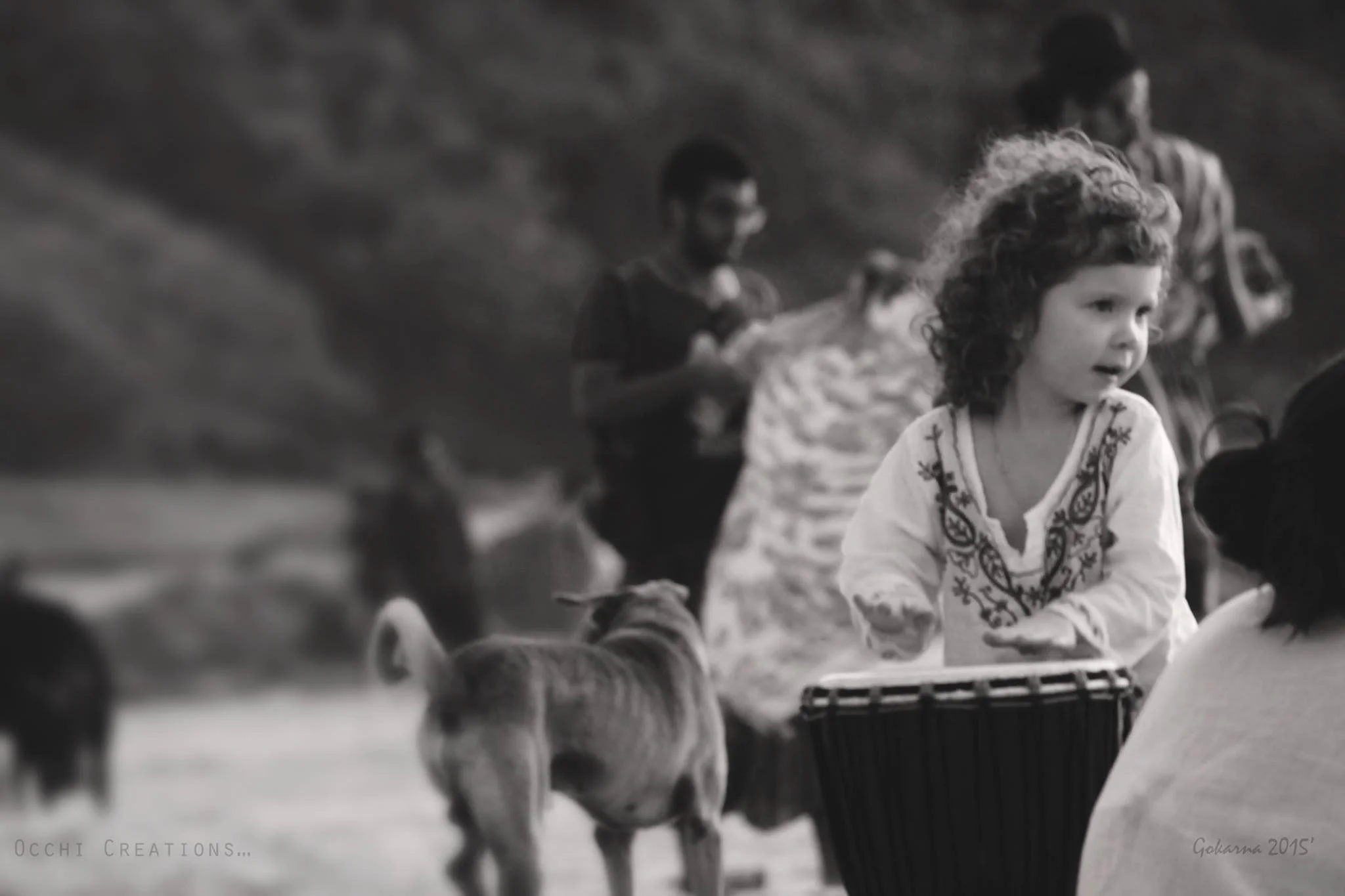A young girl with curly hair wearing a white embroidered top, standing outdoors with other children and a dog nearby. The children appear to be engaged in an activity, with some holding objects, and there is a natural outdoor setting in the backgroun