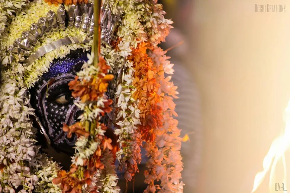 Close-up of a person with traditional Indian face paint and jewelry, adorned with orange and white flower garlands.