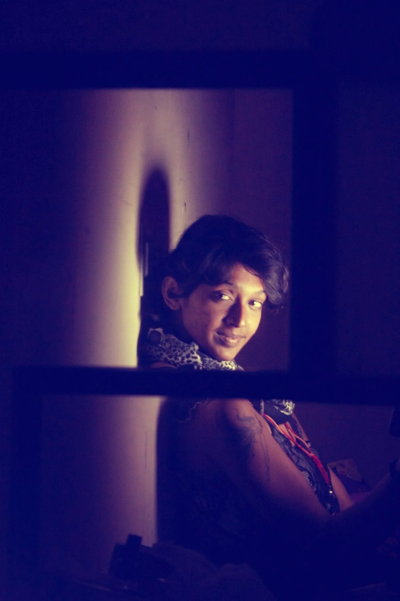 A young boy with dark hair and wearing a leopard-print shirt, seen through a narrow gap, looking toward the camera with a slight smile in a dimly lit room.