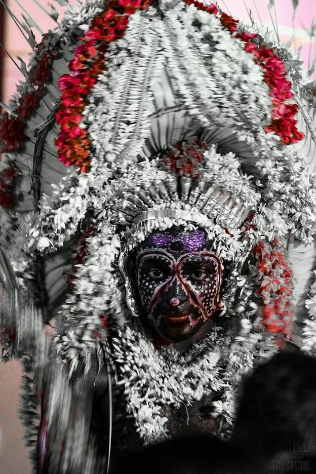 A person wearing an elaborate traditional headdress decorated with white and red flowers, with face painted in intricate black and purple designs for a cultural celebration.