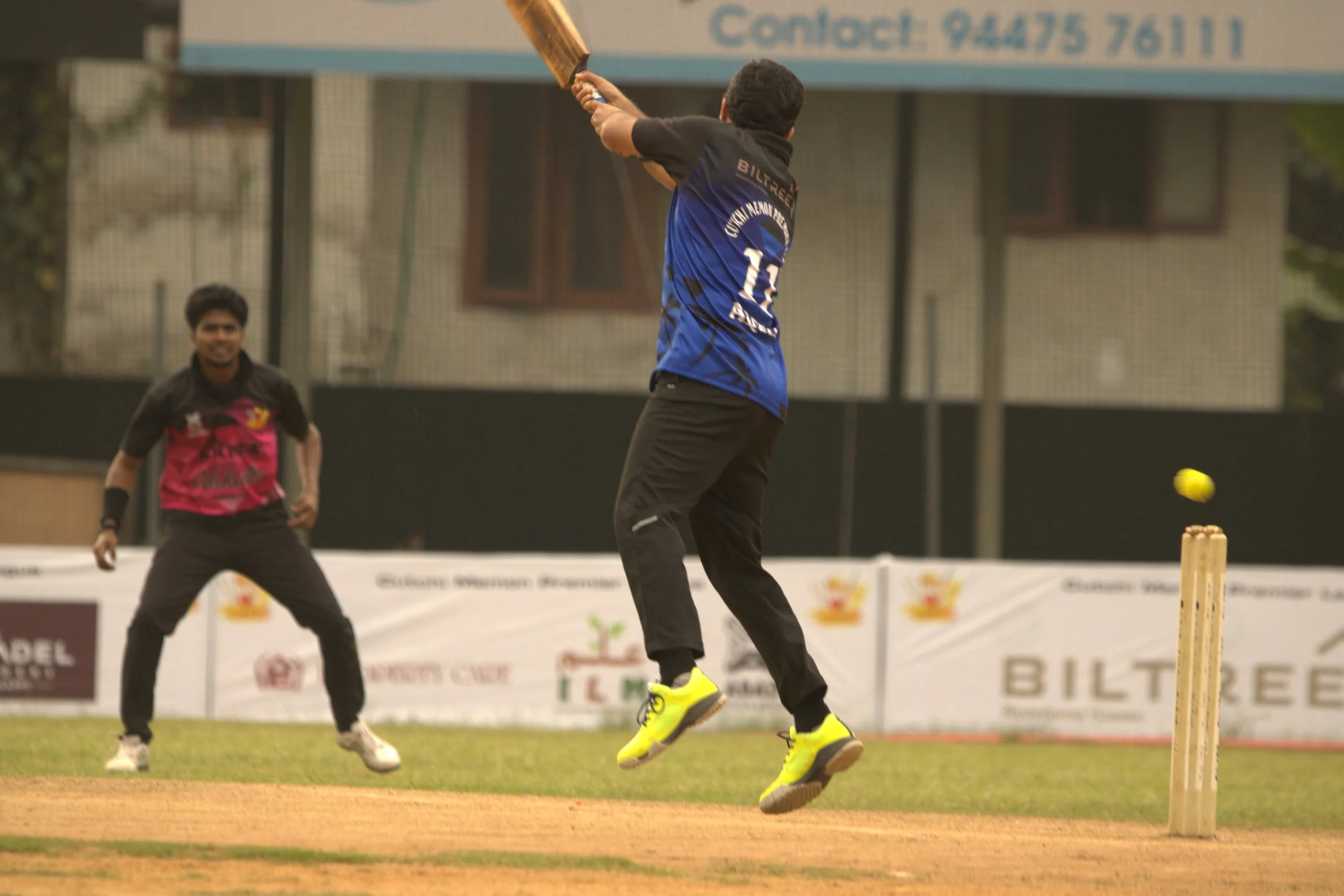 A cricket match with a player attempting a shot at the ball, while a wicketkeeper stands behind. The player is mid-air, wearing bright yellow shoes, and the ball is approaching the stumps.