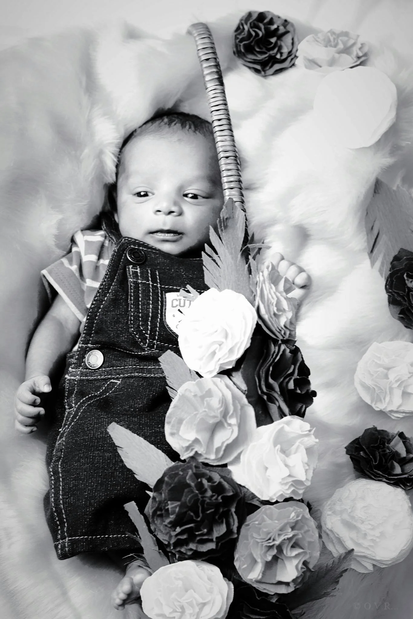 A baby lying on a soft surface surrounded by paper flowers, looking to the side. The baby is dressed in denim overalls and a striped shirt.