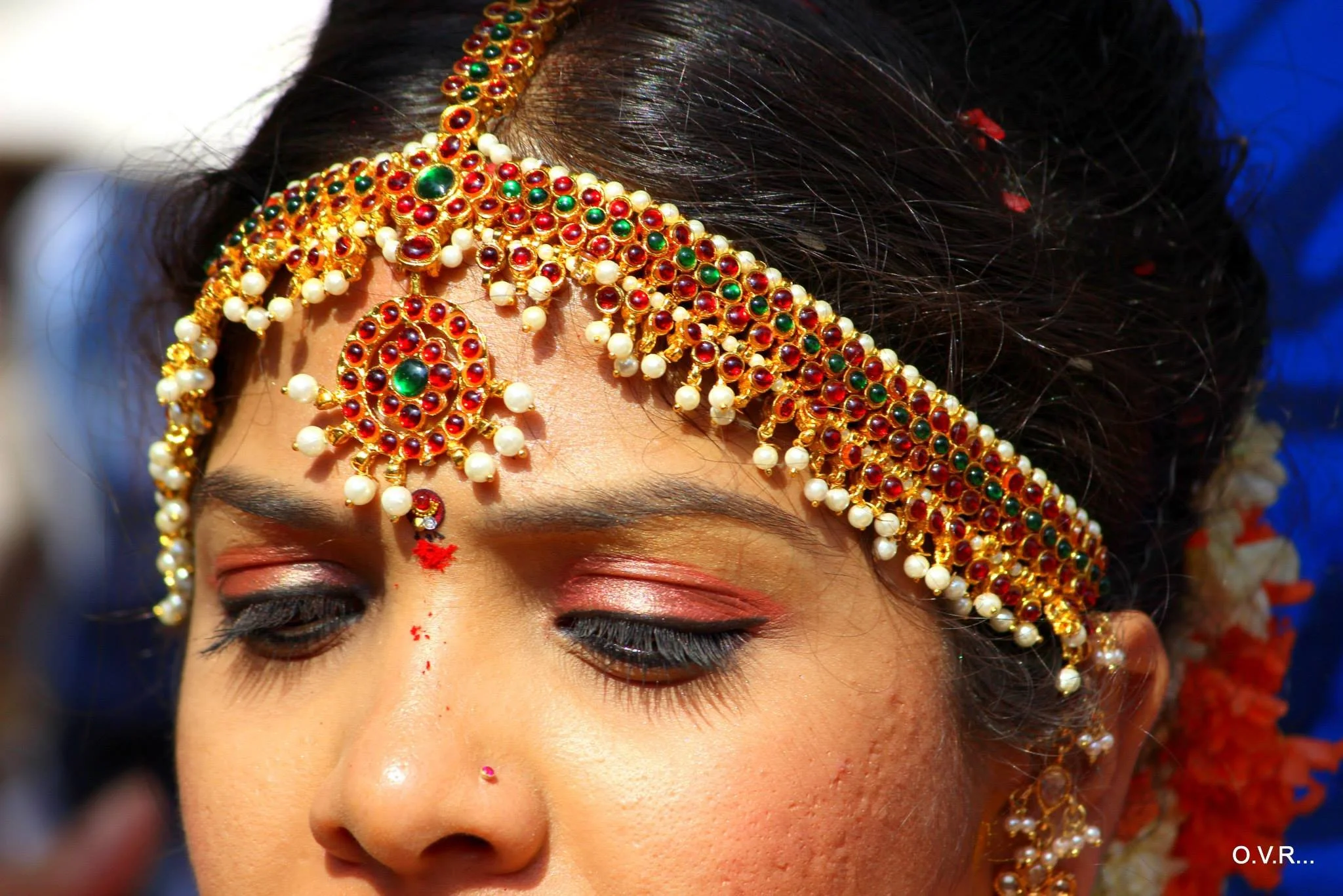 Close-up of a woman wearing traditional Indian jewelry, including a large, ornate gold and pearl headpiece with red and green gemstones, and matching earrings
