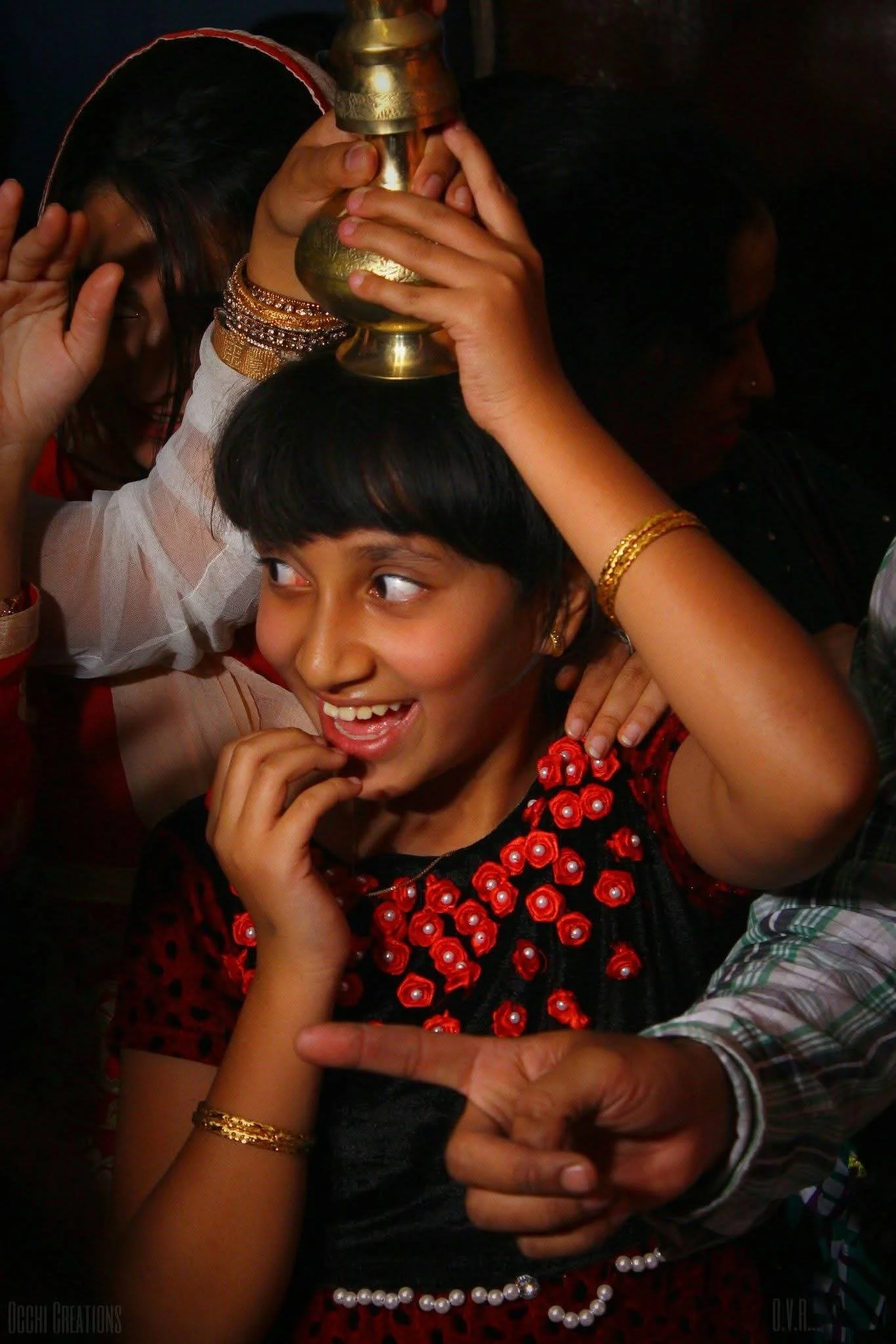 A young girl wearing a black and red dress with red floral embellishments smiles happily as she is crowned with a brass cup during a celebration. Several people are around her, some touching her head and pointing at her, in a festive and joyful atmos