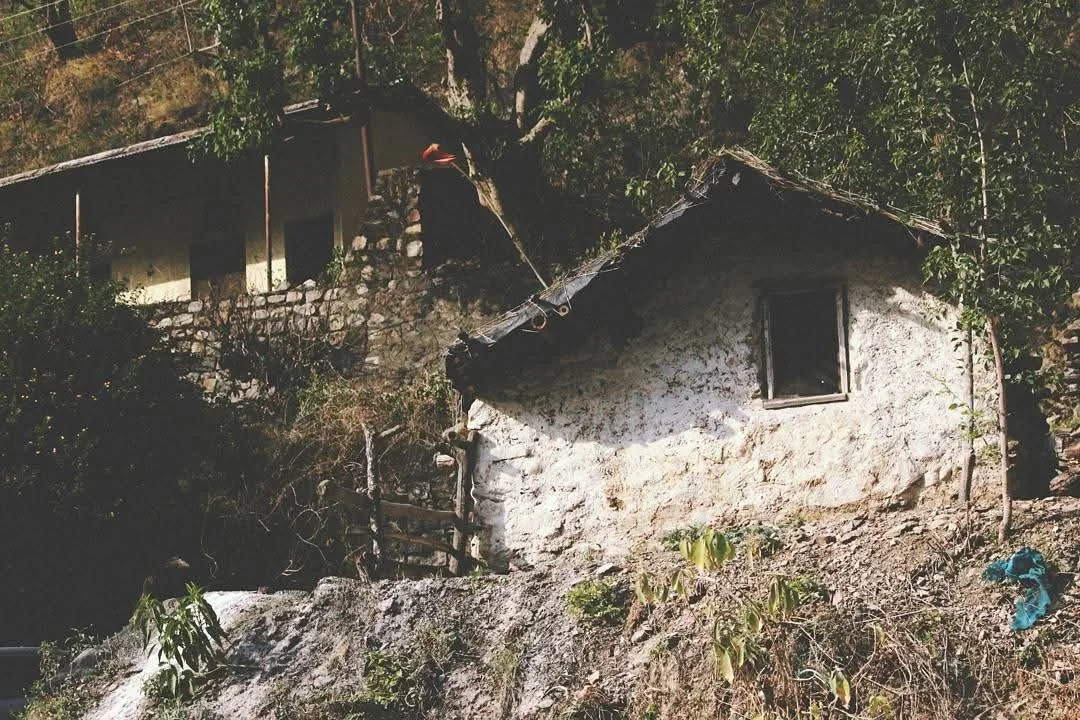 A house built into a rocky hillside, with stone walls and a window, surrounded by trees and vegetation.