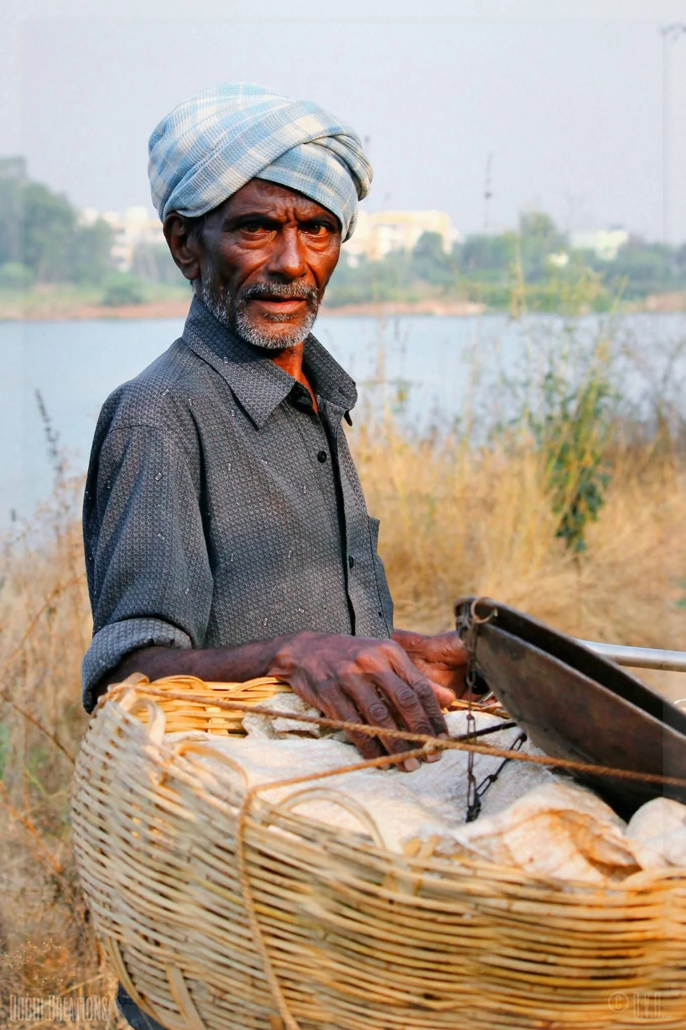 An elderly man wearing a traditional headscarf and a dark shirt holding a basket near a water body surrounded by dry grass with buildings visible in the background.