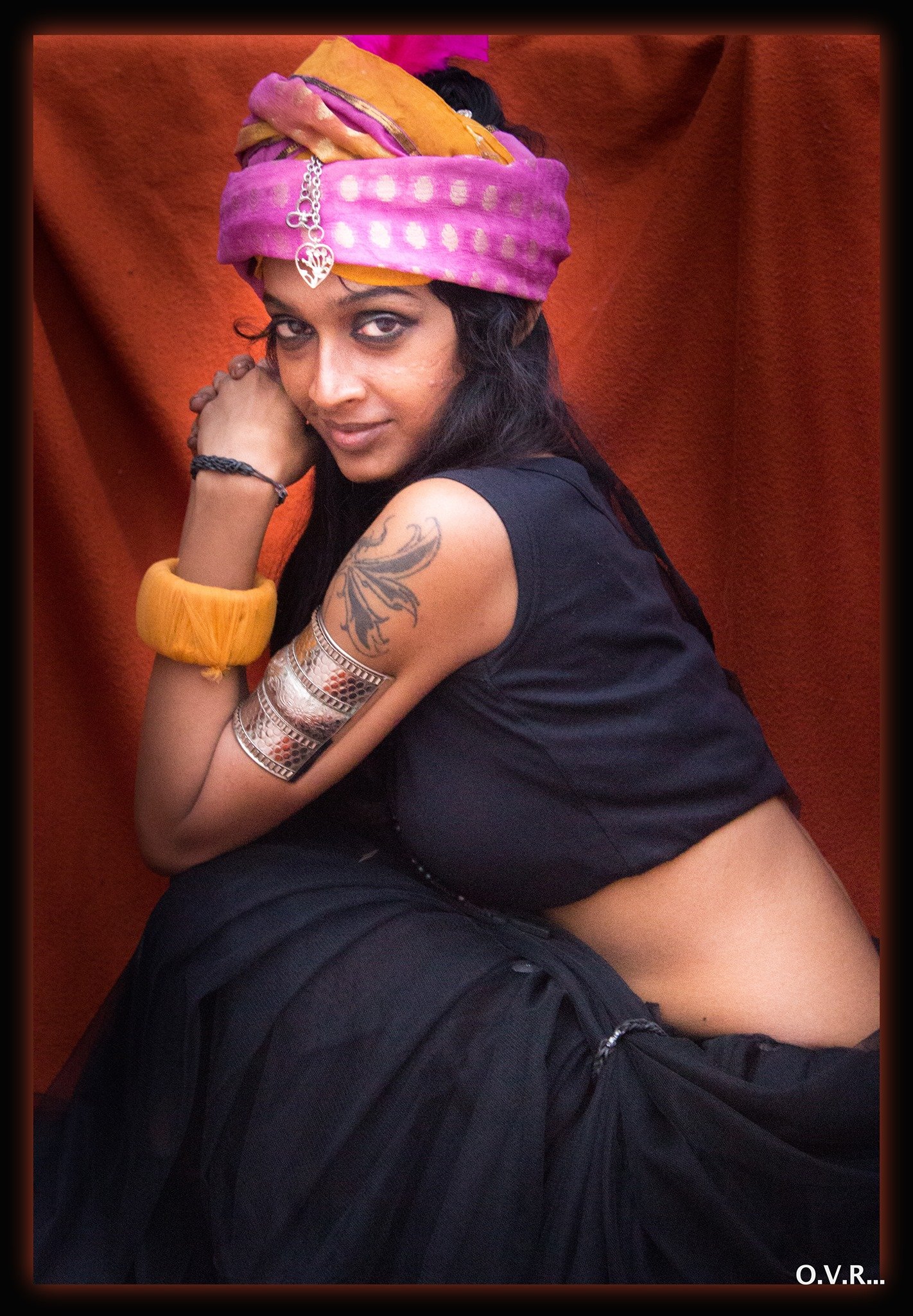 A woman with black hair, wearing a colorful headscarf, black top, and jewelry, poses against a reddish-brown backdrop. She has a tattoo on her upper arm and is leaning forward with her elbow on her knee, looking at the camera.
