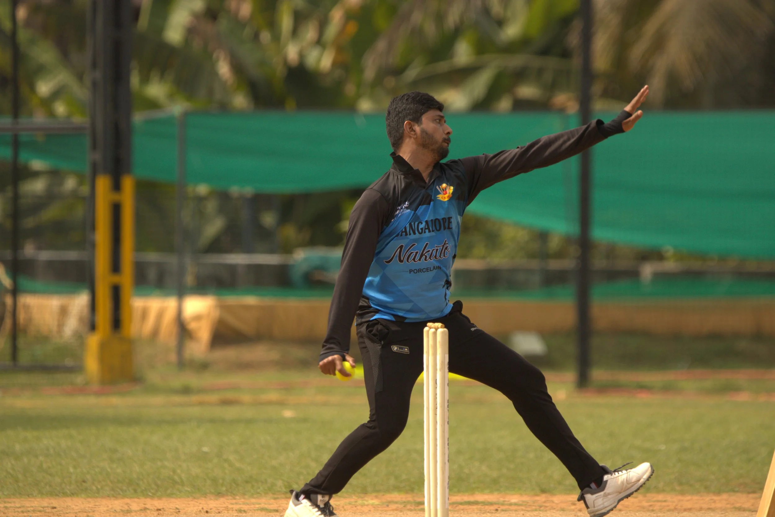 A cricket player is in the middle of a bowling action on a cricket field, wearing a black and blue jersey with the text 'Bangalore Nakato' and holding a yellow cricket ball.