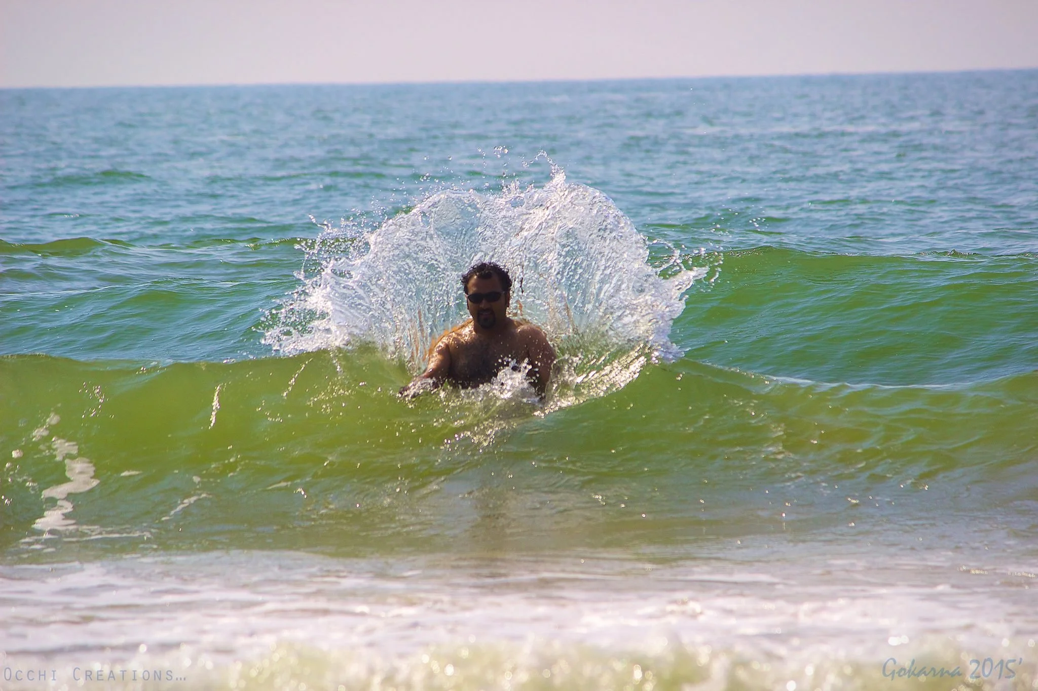 A man with dark hair and a beard, wearing sunglasses, is playing in the ocean waves, creating a splash with his hands.