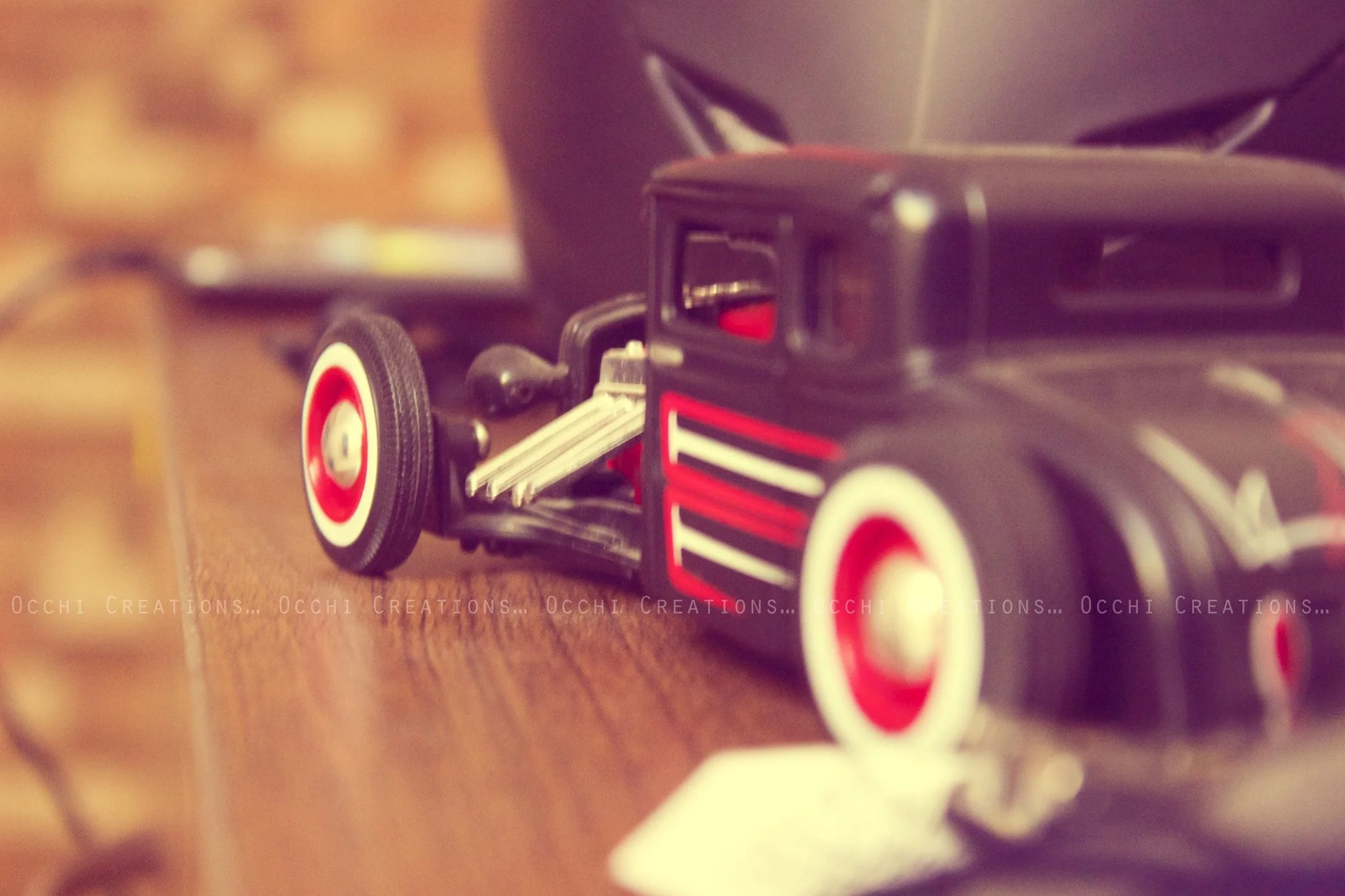 A black toy car with red and white stripes and a white license plate, situated on a wooden surface. In the background, a blurred fork and a black hat are visible.