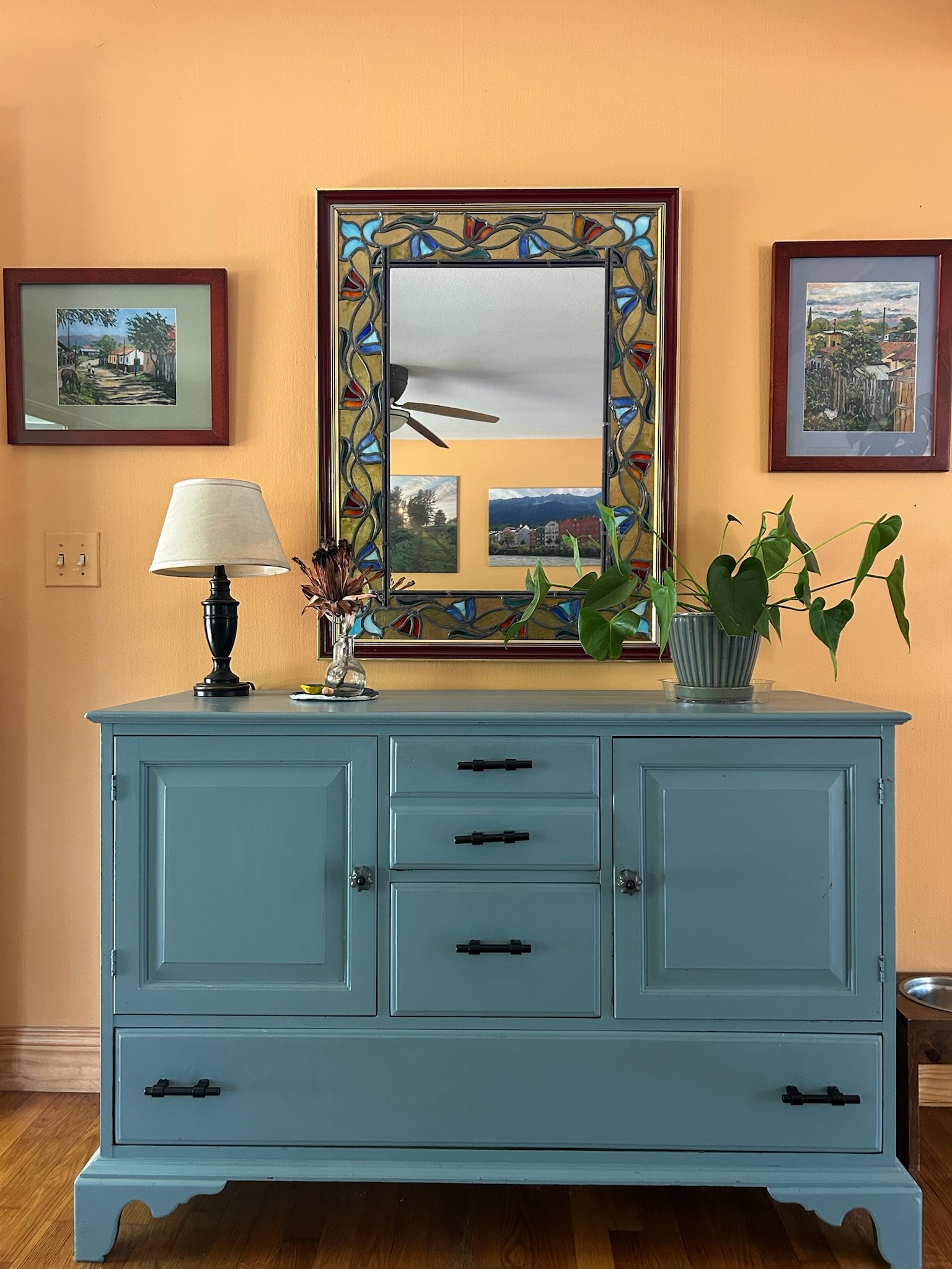 A blue dresser set against a yellow wall with framed images and decorative items on it. An example of a room organized by Creating Joyful Spaces