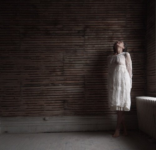 Woman in a white lace dress standing against a dark wooden wall, looking upward with her eyes closed.