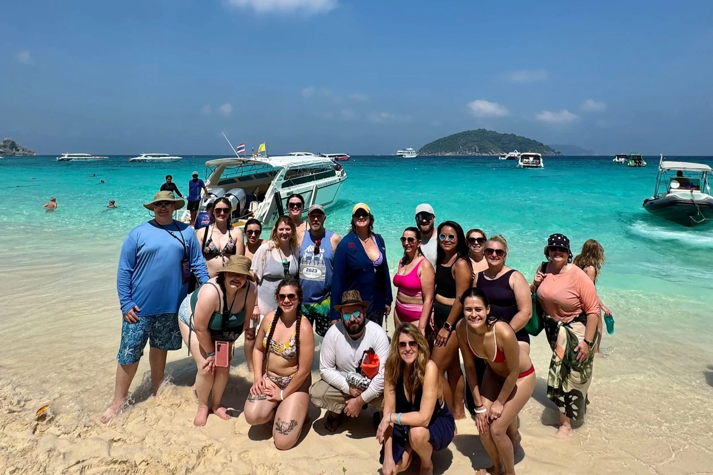 Group of people in swimsuits and casual summer clothes standing on a sandy beach with turquoise water, boats in the background, and a small island in the distance.