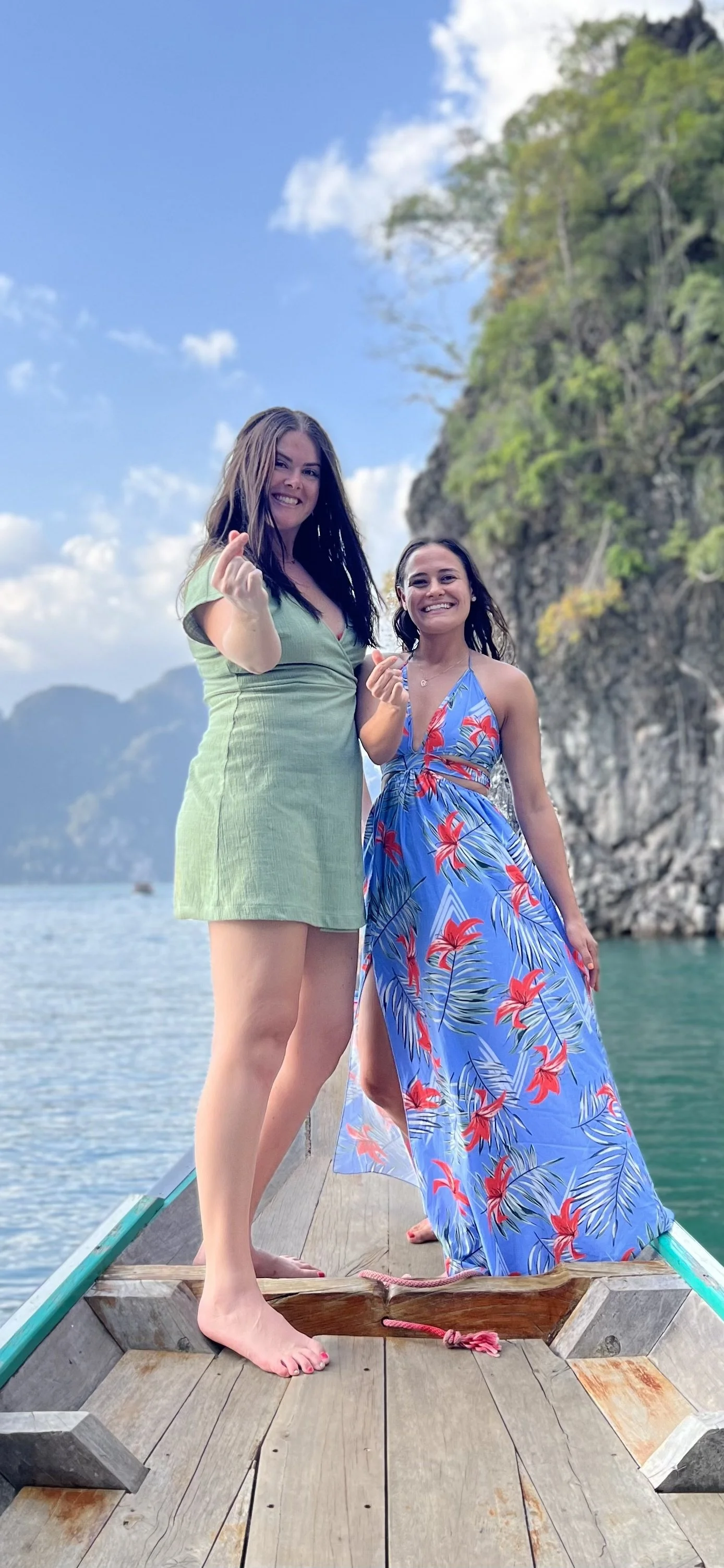 Two women standing on a wooden boat at a lake with steep, forested cliffs in the background, smiling and making a hand gesture.