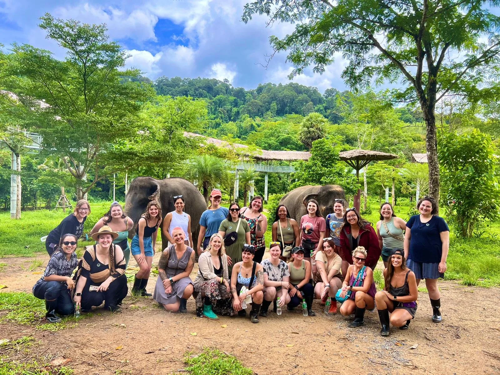 Group of diverse women outdoors at a zoo or wildlife sanctuary, with elephants and lush greenery in the background, under a bright blue sky.