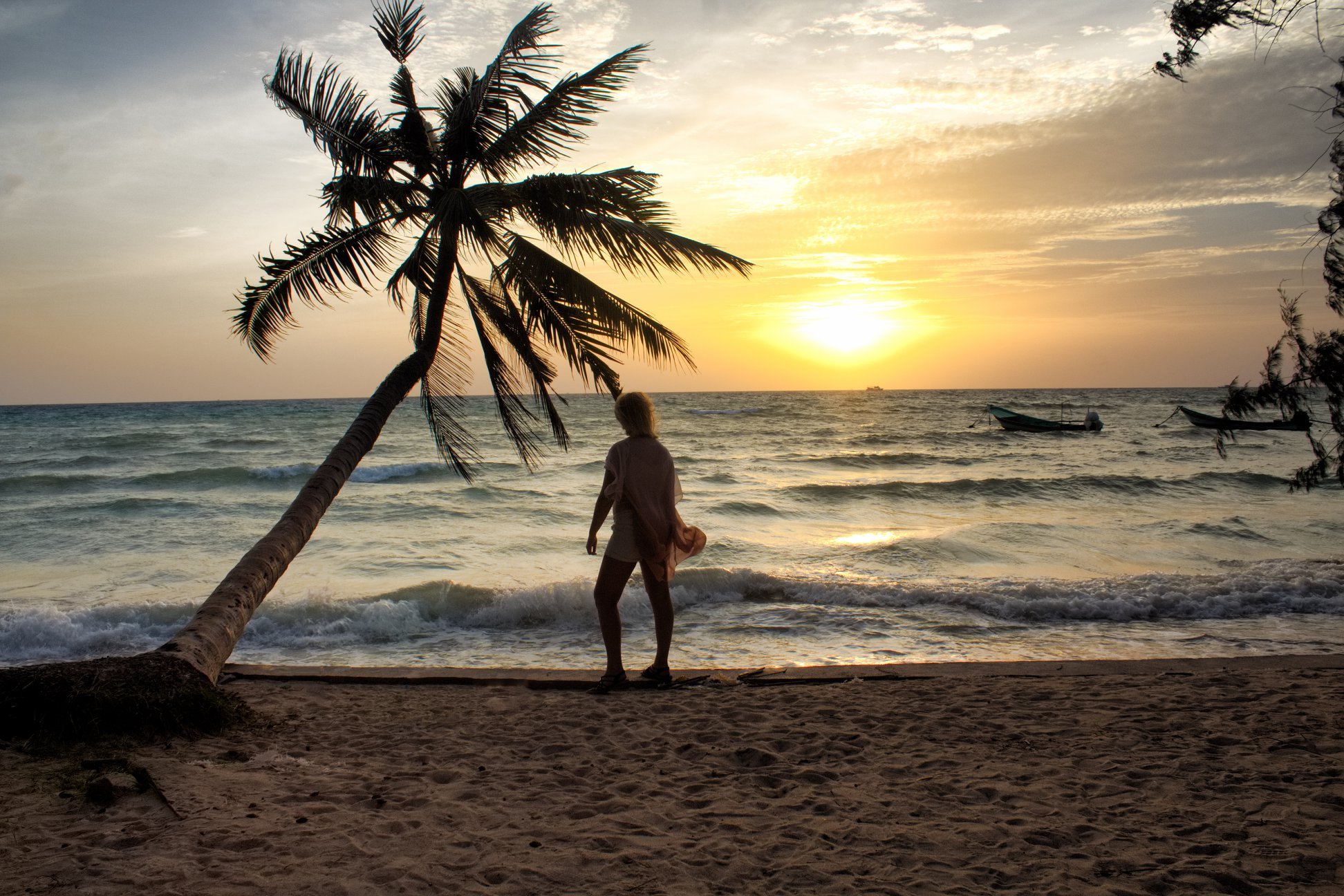 A person standing on a sandy beach under a leaning palm tree during sunset, with boats floating on the ocean in the background.