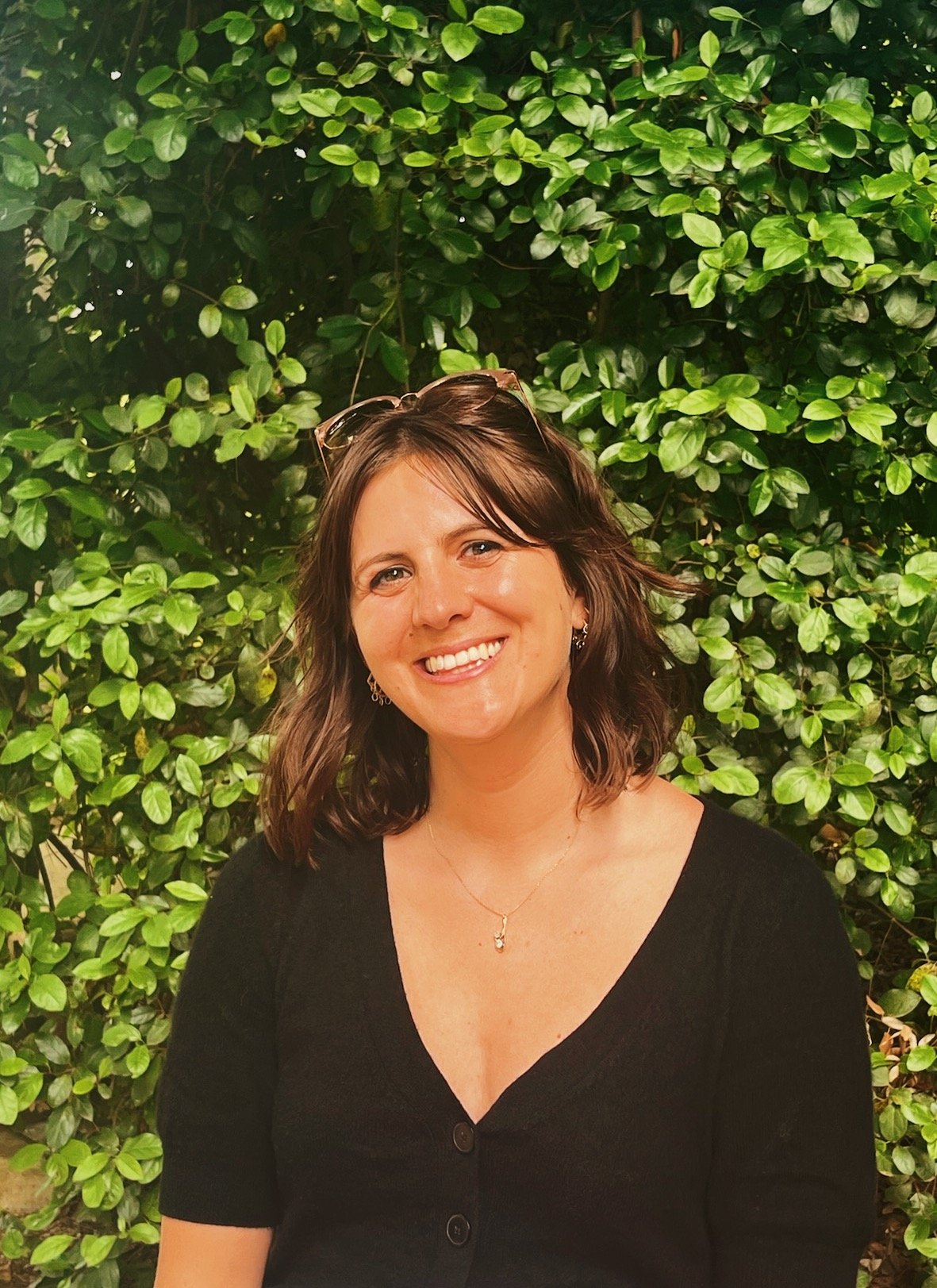 A woman with brown hair smiling, wearing a black top, earrings, a necklace, and sunglasses on her head, standing in front of a lush green leafy background.
