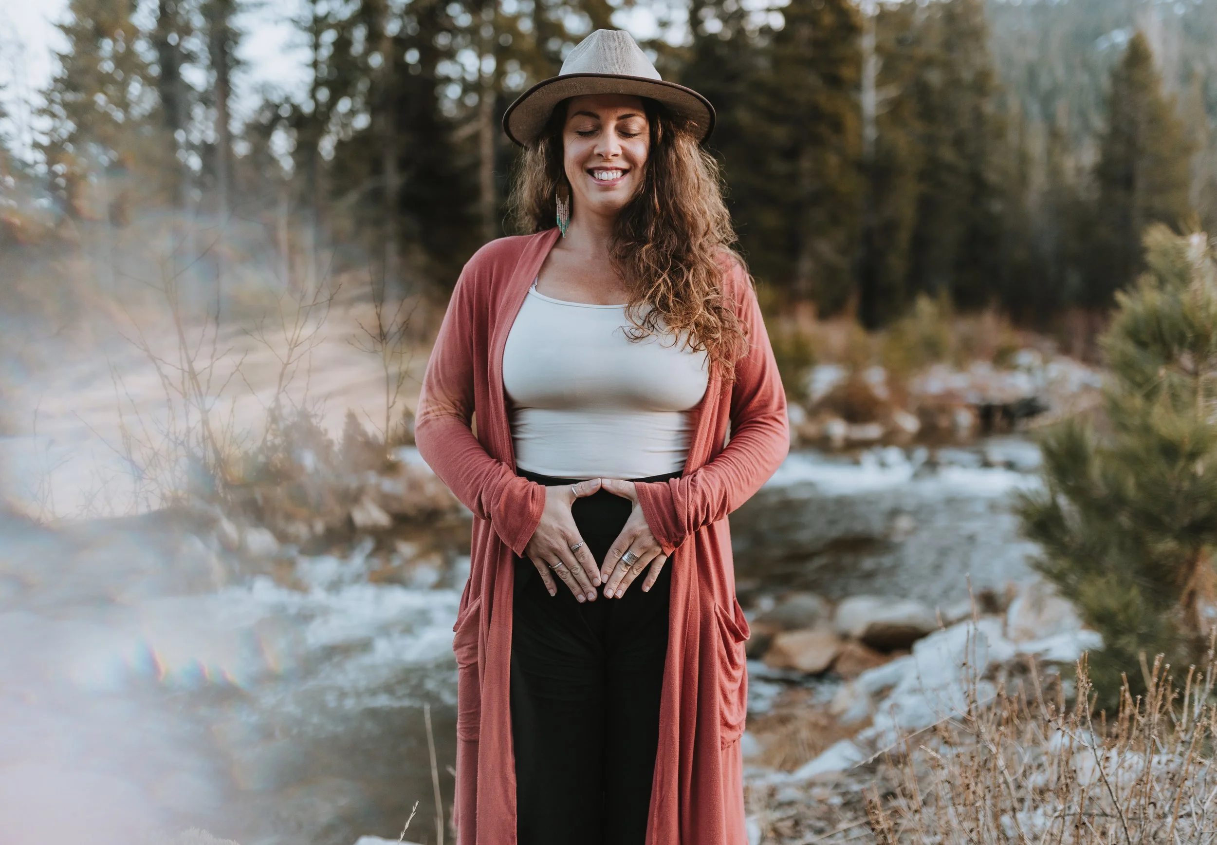 A woman with long, curly hair wearing a wide-brimmed hat, white top, pinkish-brown open-front cardigan, and black pants, standing outdoors near a stream, smiling with eyes closed, making a heart shape with her hands over her stomach.