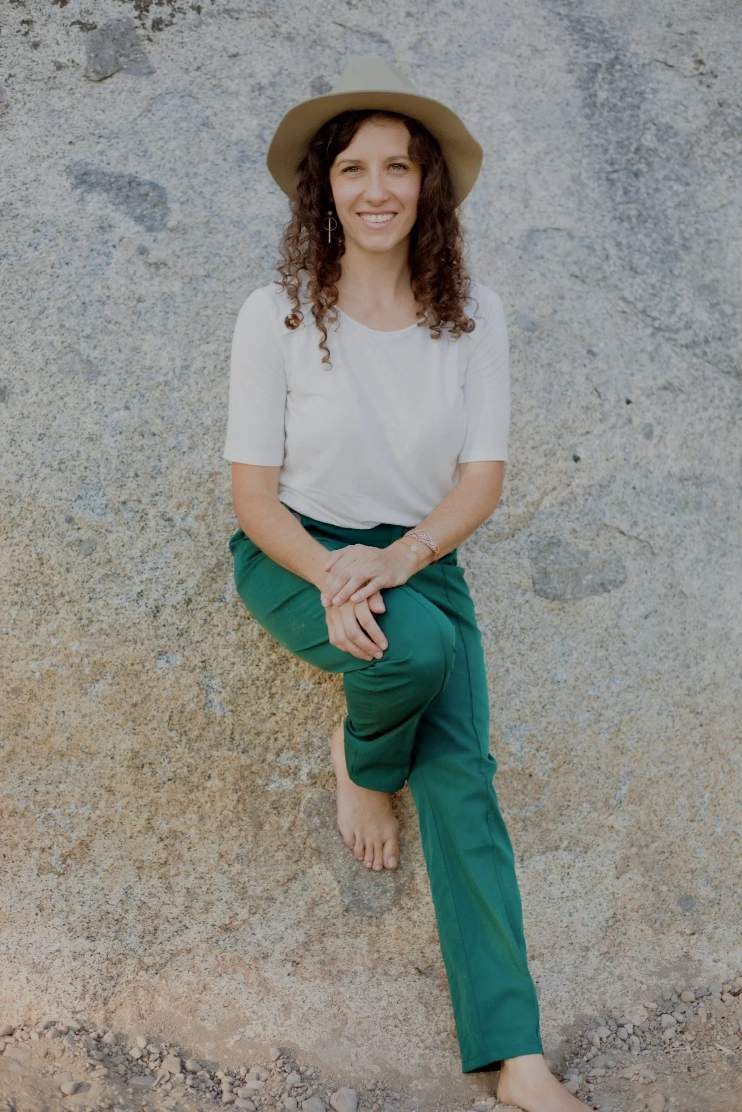 A woman sitting against a rock wall, wearing a white t-shirt, green pants, a beige hat, and earrings, with a smiling expression.