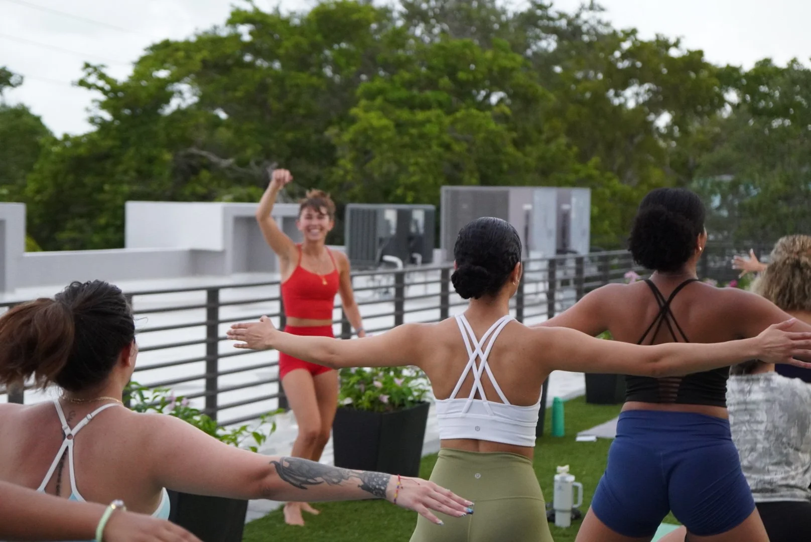 Group of women in Miami participating in outdoor Pilates or fitness class on rooftop with green trees in background, instructor leading session, women in colorful athletic wear with arms extended.