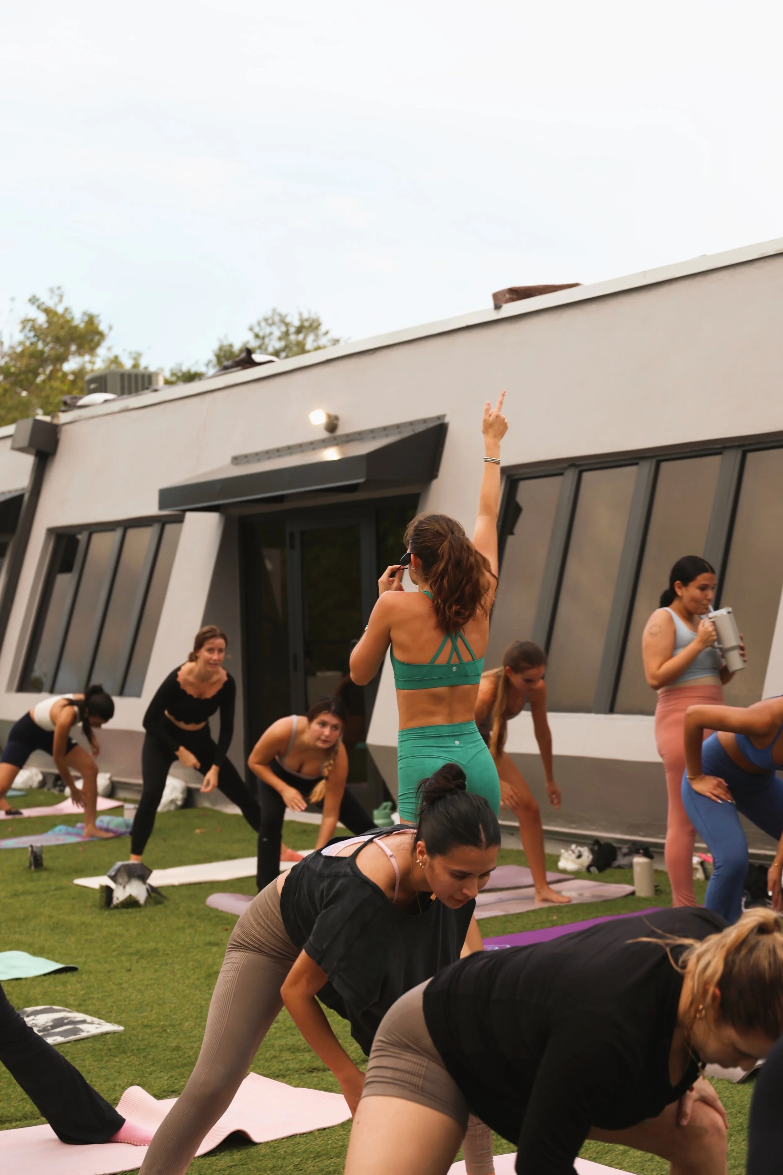 Women participating in an outdoor yoga class on grass in front of a modern building in Miami, with one woman in green workout attire standing and making a victory sign while talking on her phone.