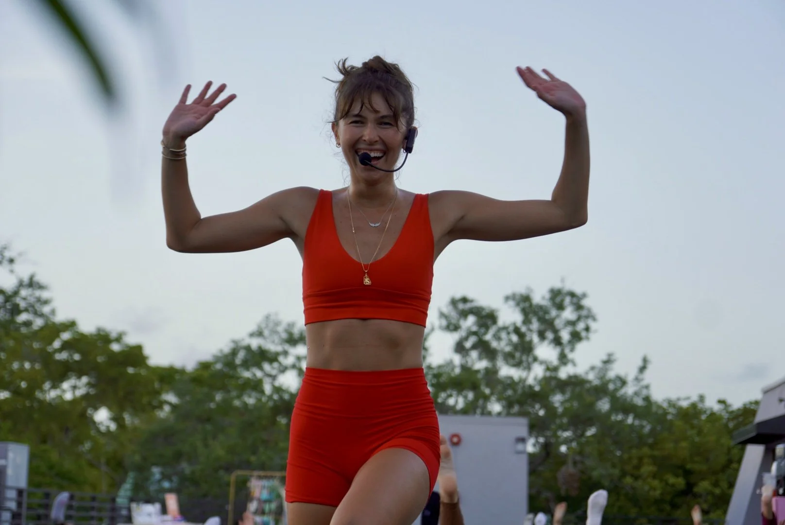 Pilates instructor in red workout outfit in Miami smiling and raising her arms outdoors, wearing a headset microphone, with trees and a cloudy sky in the background.