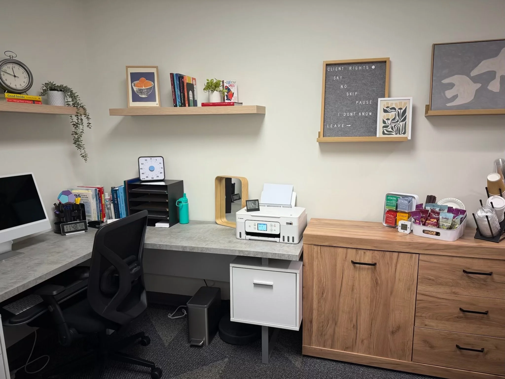 Office workspace with a desk, computer, books, printer, and storage cabinet; decorative shelves and wall art.