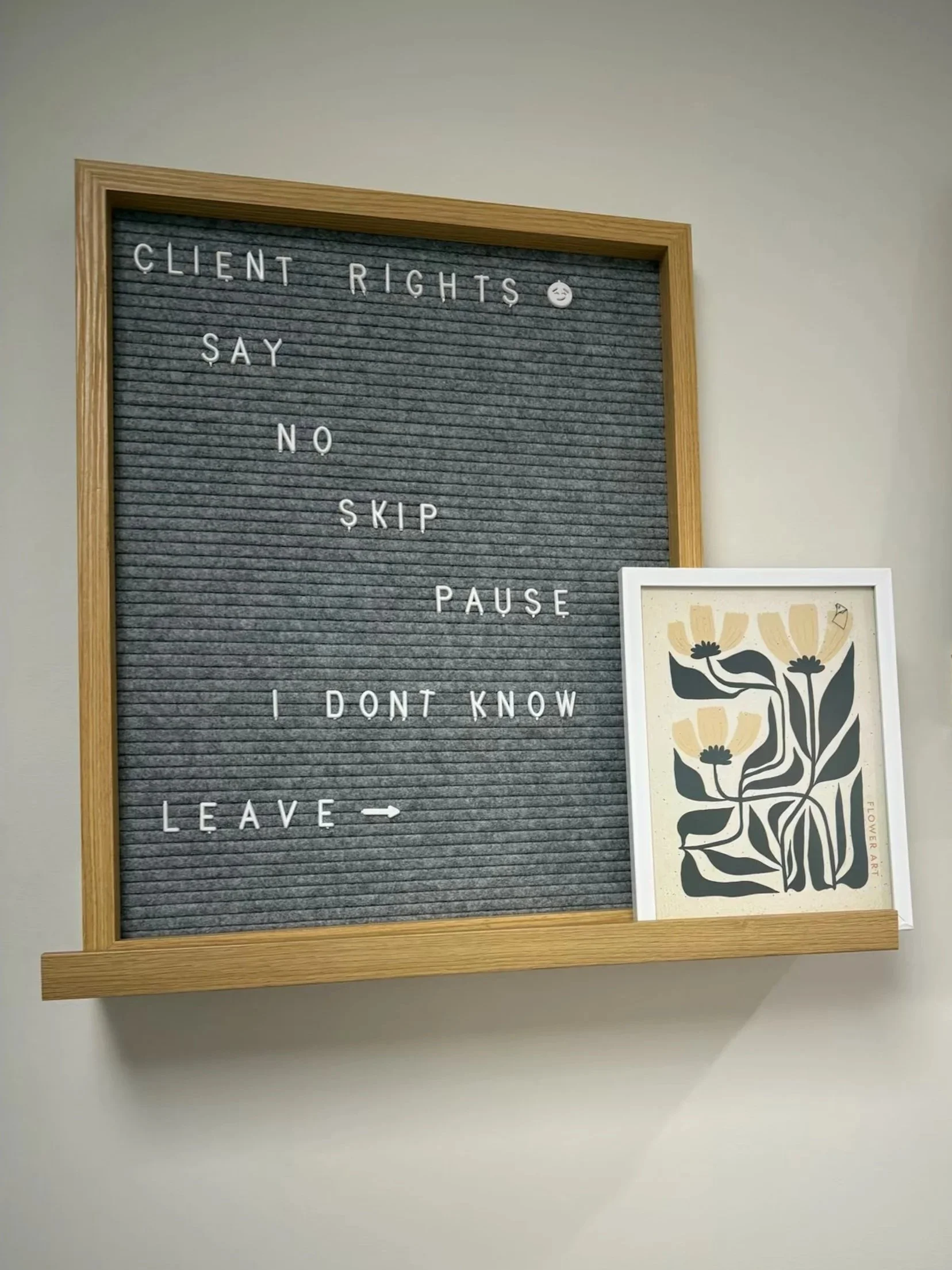 A felt letter board with the message: "Client rights say no skip pause I don't know leave" and a small smiling face emoji. Beside it, a framed flower art print with black and beige flowers and leaves.