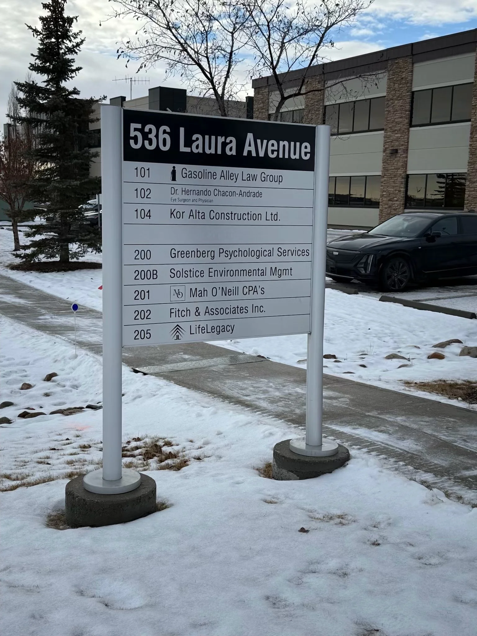 Building directory sign on Laura Avenue street with parking lot and snow-covered ground in the foreground, trees, a modern building, and a black car in the background.