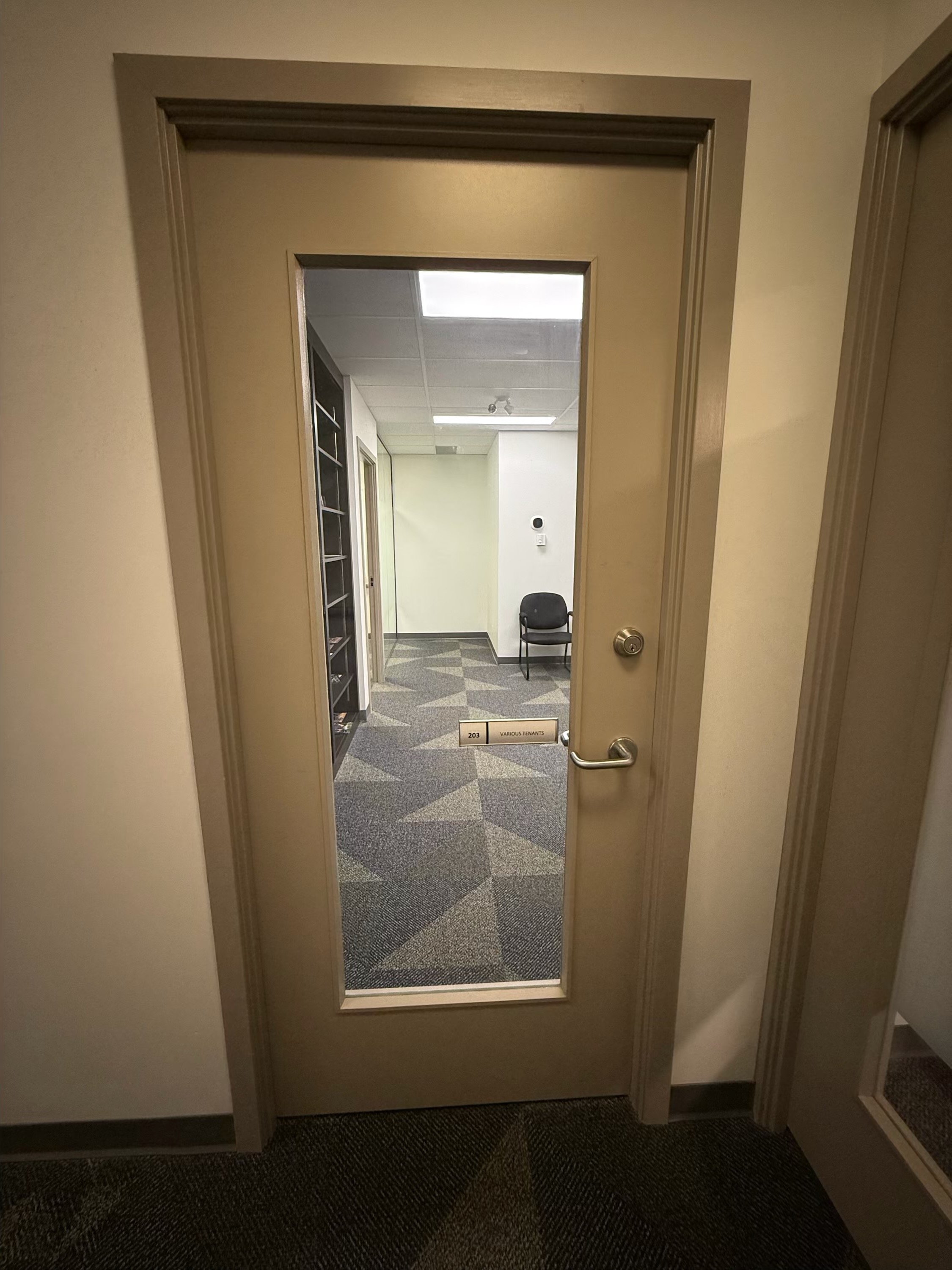 Interior view through a glass door into a small office or waiting area with black chair, bookshelves, and patterned carpet.