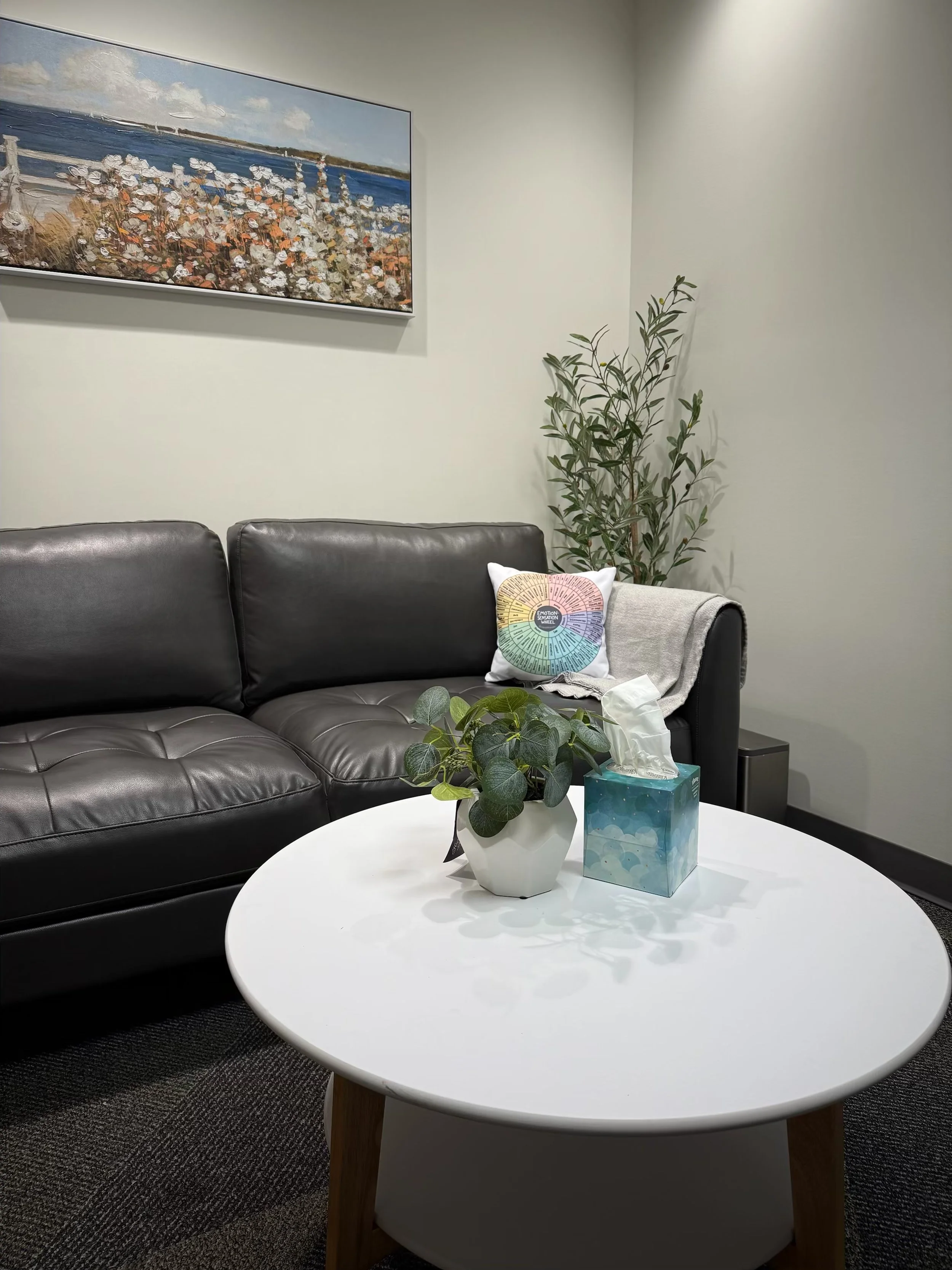A waiting room with a black leather couch, a white round table with a small potted plant and a tissue box, a colorful cushion on the couch, a beige throw blanket, a tall plant in the corner, and a landscape painting on the wall.