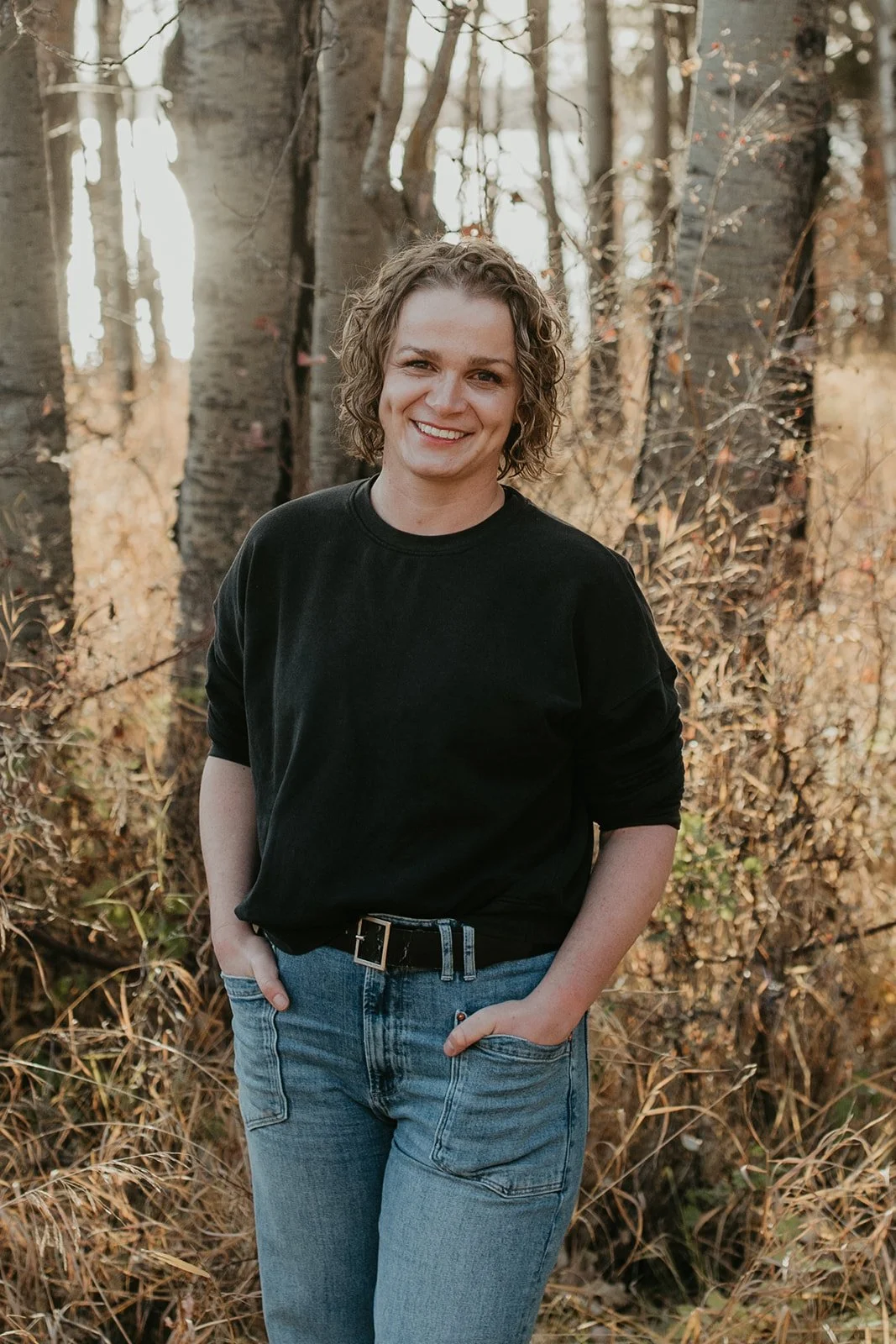 A woman with curly hair wearing a black shirt and blue jeans standing outdoors in a wooded area with brown and orange foliage.