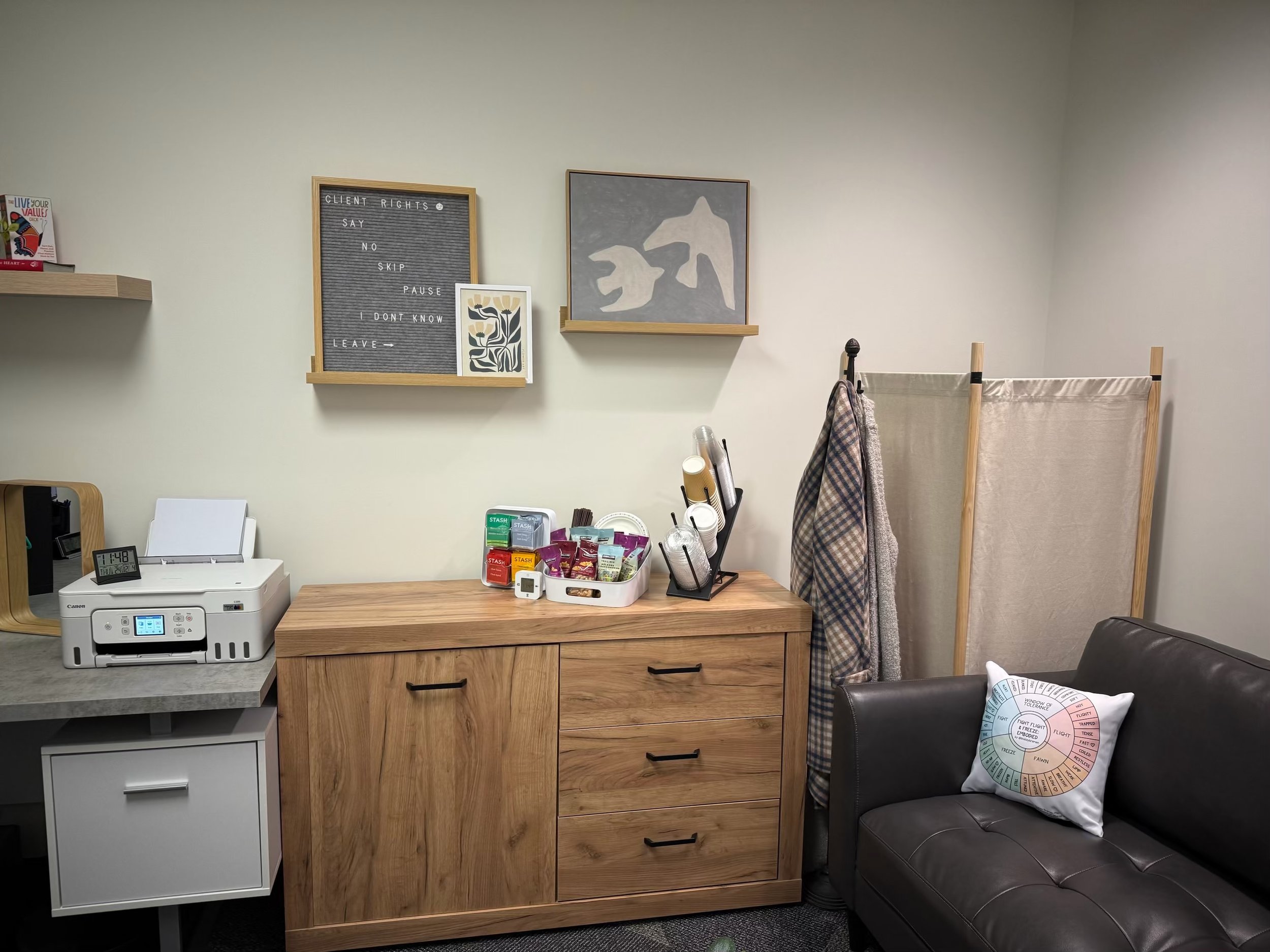 Office or waiting room with a wooden cabinet, a black leather couch with a colorful circular pillow, a white printer, framed artwork, a wall letter board with a message, and a fabric privacy screen.