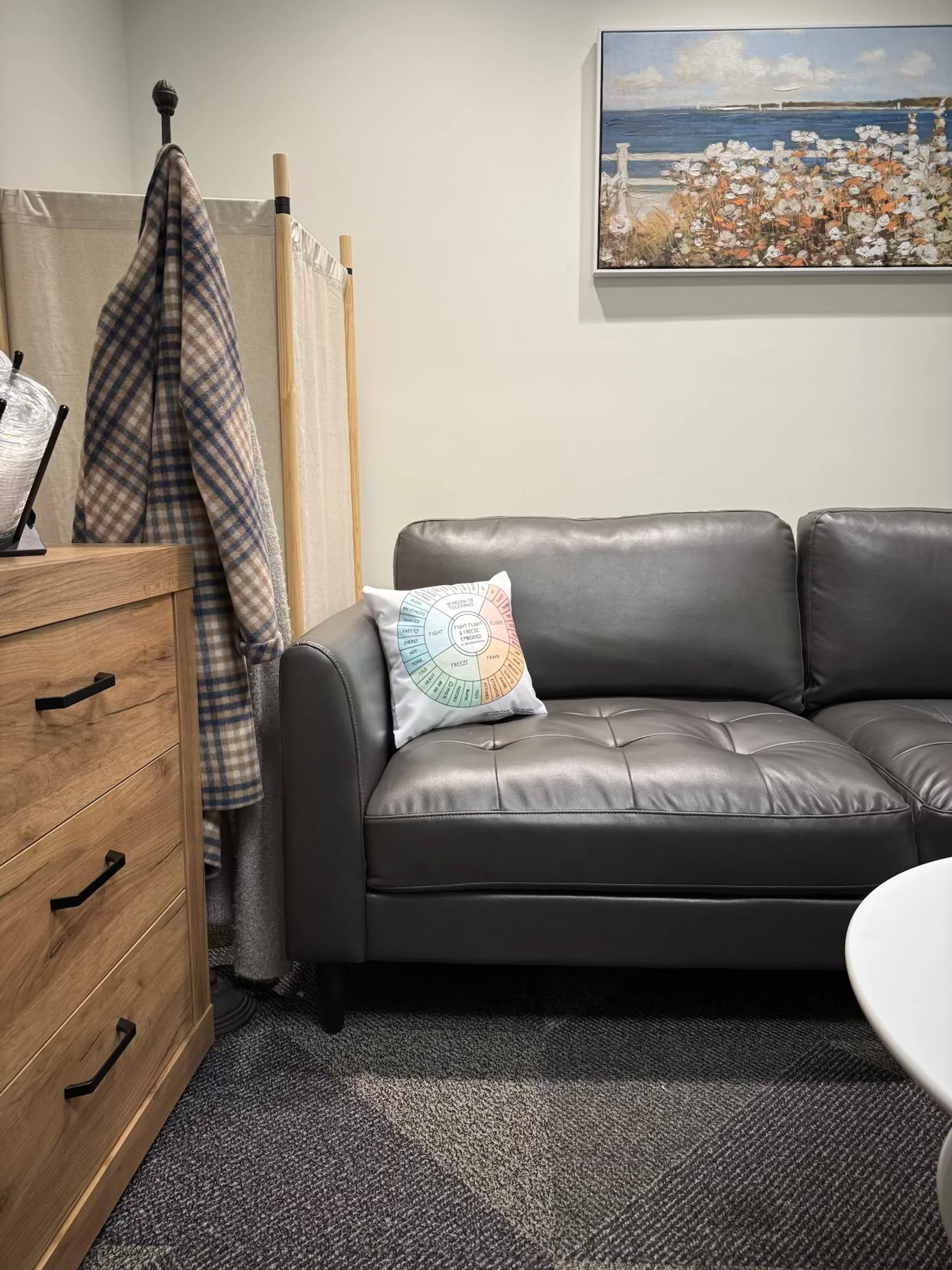 A living room corner with a gray leather couch, a colorful round pillow, a wooden dresser, and a painting of a beach scene on the wall.
