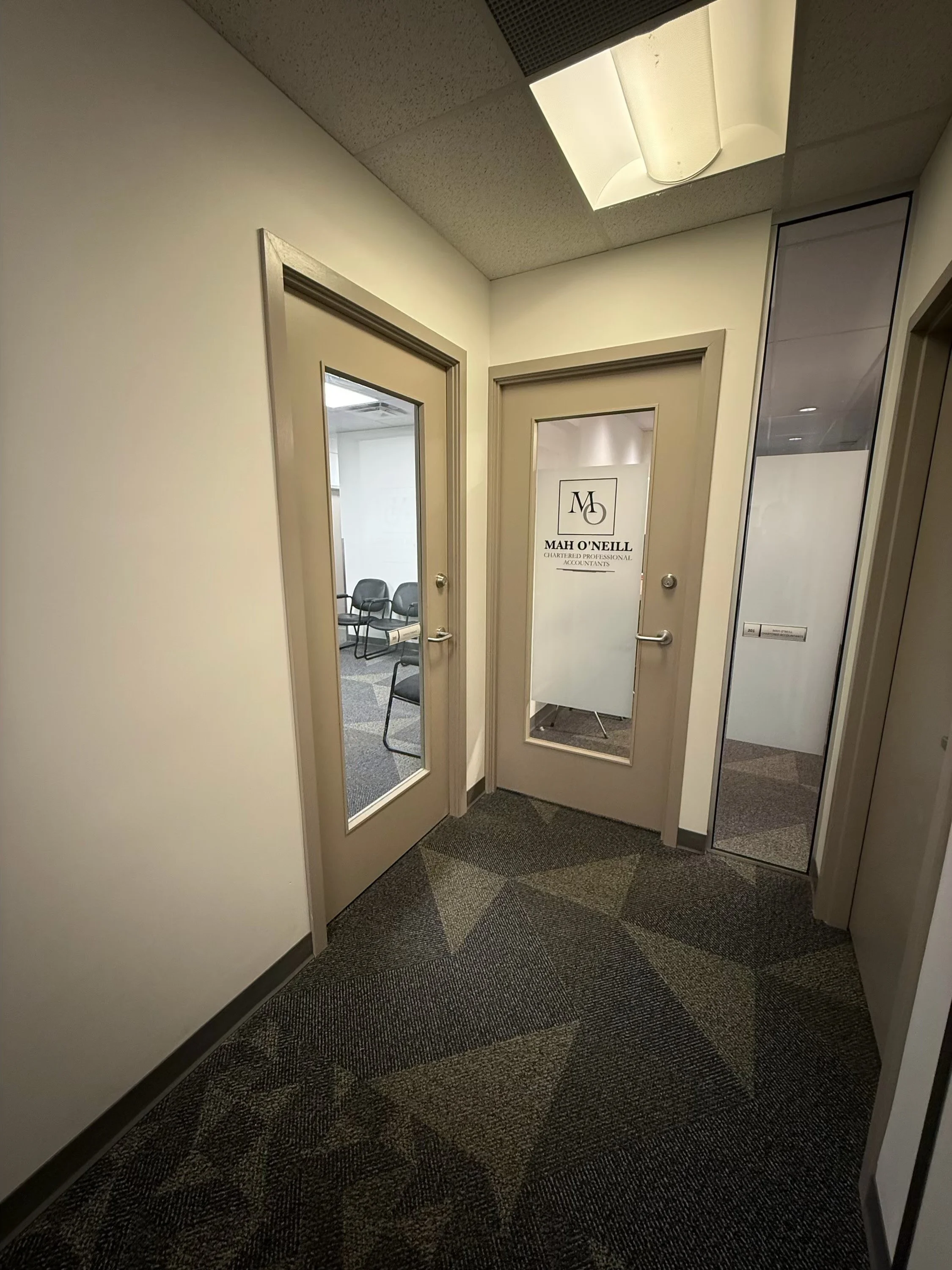 A hallway in an office building with two beige doors, one with a clear window and one with frosted glass, both leading to separate rooms. The door on the frosted glass has the logo and name of Mah O'Neill, Chartered Professional Accountants. There is