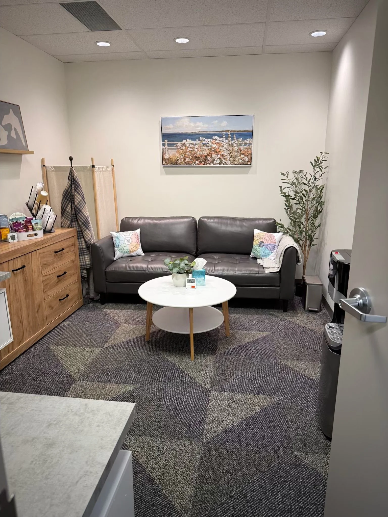 A cozy waiting room with a dark gray leather sofa, a white oval coffee table with wooden legs, and a wooden cabinet on the left side. The room has a gray patterned carpet, a tall potted plant on the right, and a framed painting of a beach scene with 