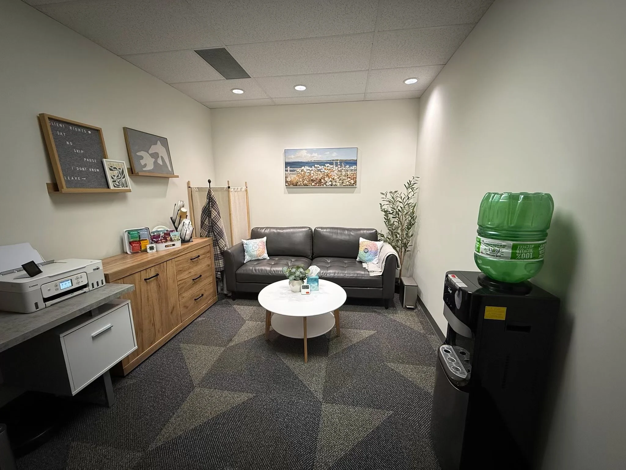 Office lounge with a black leather sofa, a white round coffee table, a wooden cabinet, a potted plant, a water dispenser, and framed wall art.