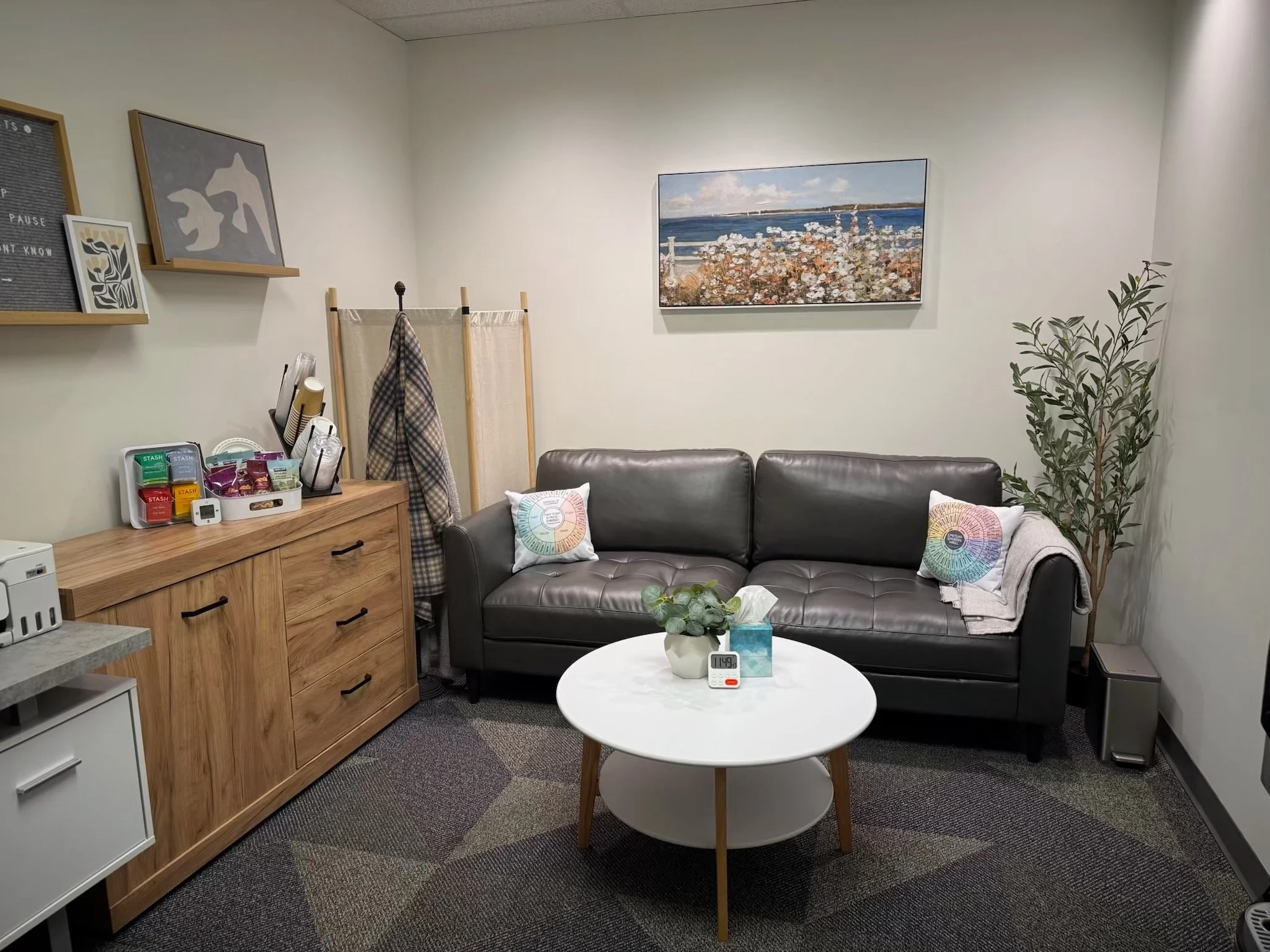 A cozy office or waiting room with a black leather couch, white round coffee table, and a gray carpet with geometric patterns. Decor includes pillows, a plant in a pot, artwork on the walls, a wooden sideboard with snacks and accessories, and a beige