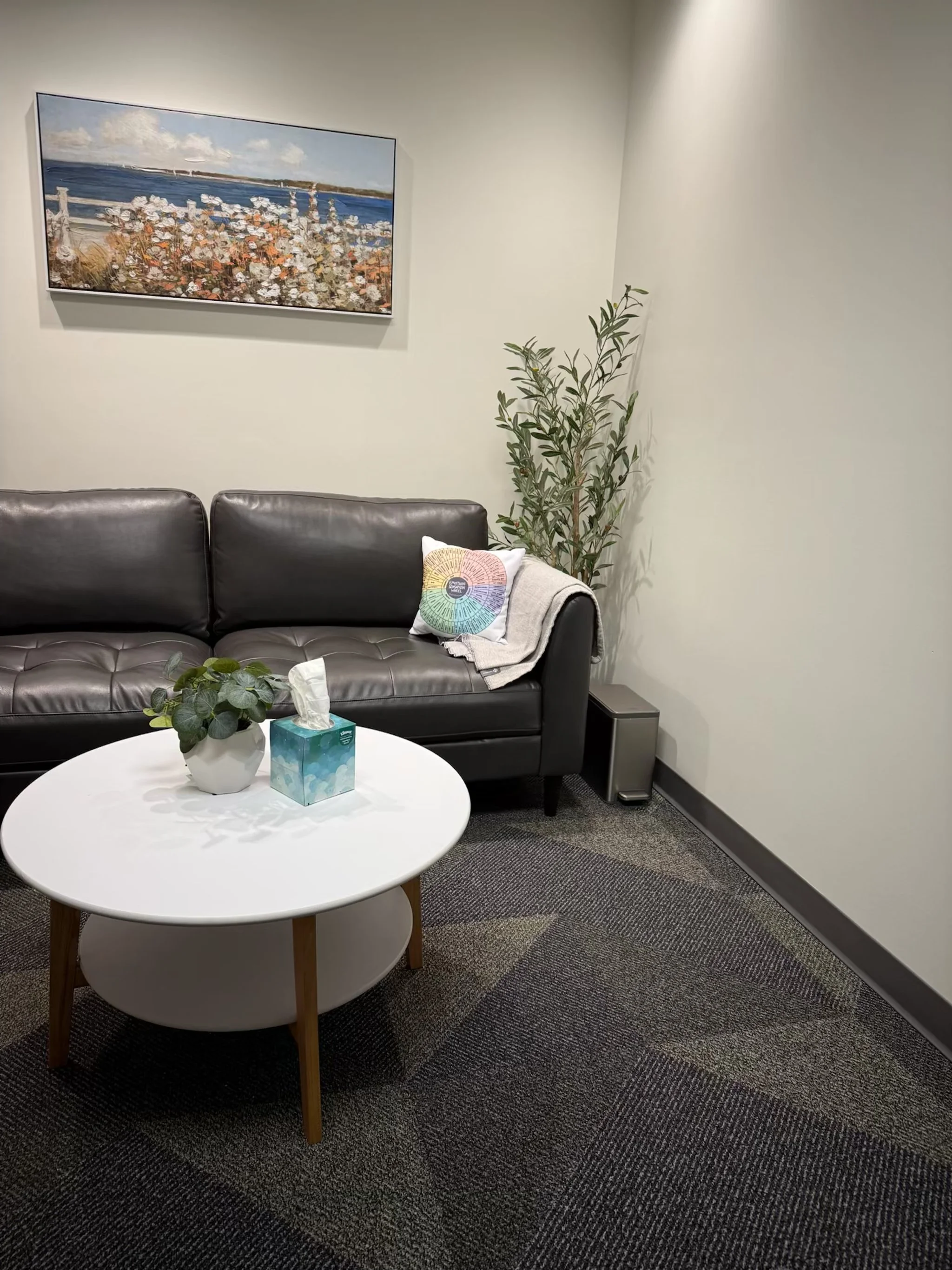 A cozy waiting room with a black leather couch, a white round coffee table with a tissue box and a potted plant, a tall potted plant behind the couch, a framed landscape painting on the wall, and a colorful pillow on the sofa.