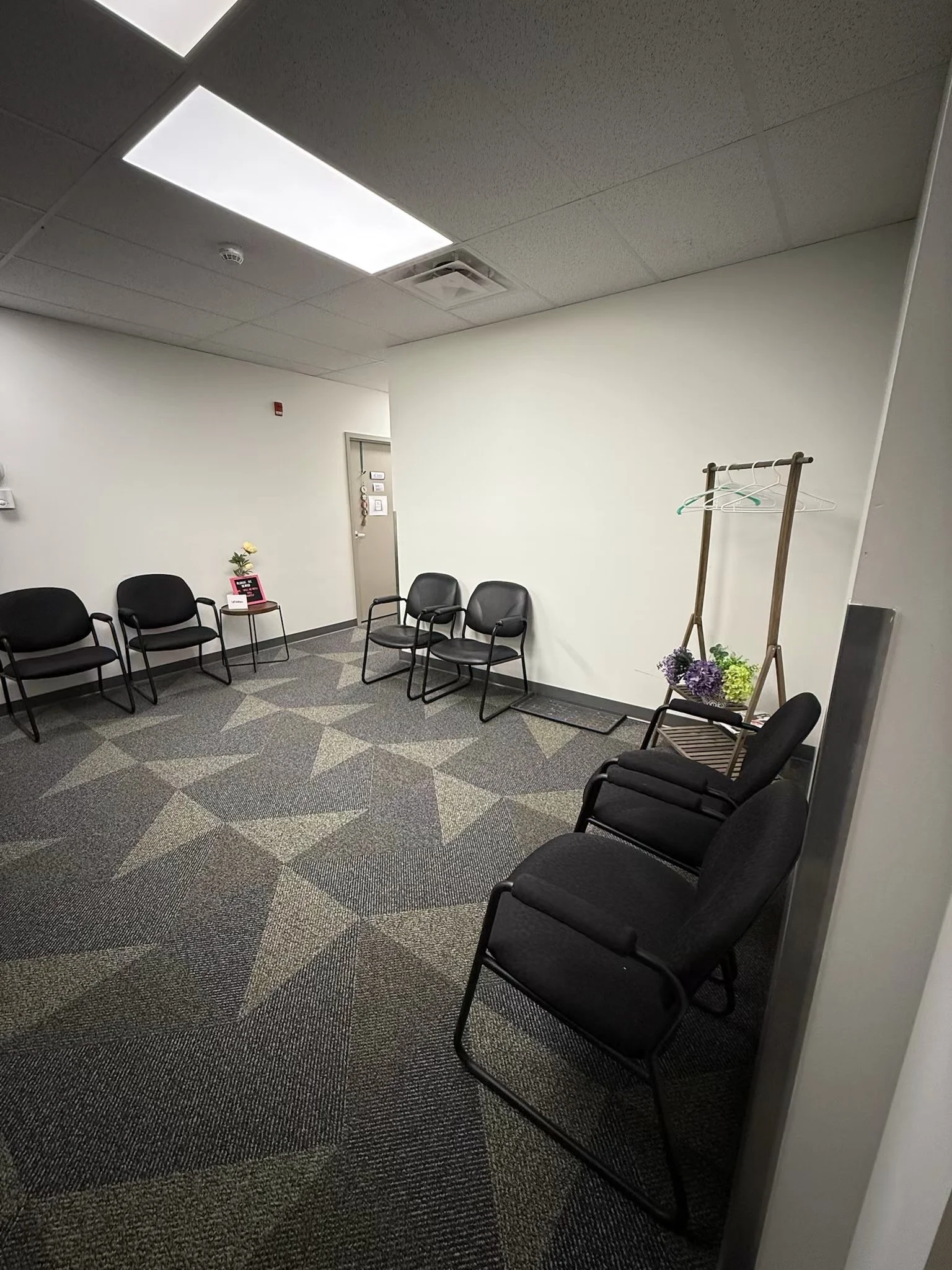 Empty waiting room with black chairs arranged along the walls, a small table with flowers and a pink sign, and a coat rack holding some hangers and flowers, with a plain white wall and ceiling tiles in the background.