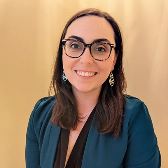 Mallori is smiling against a yellow background, wearing black glasses and a blue blazer and black blouse. 