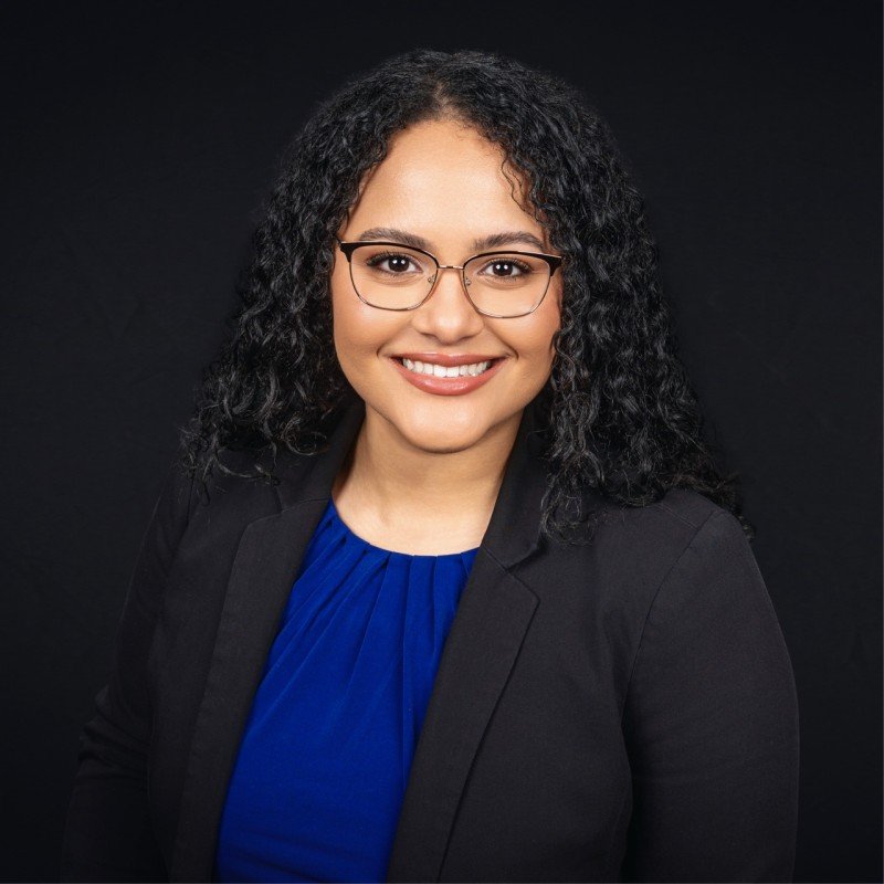 Arianna Greene is smiling against a black background, wearing glasses and a black blazer over a blue top.