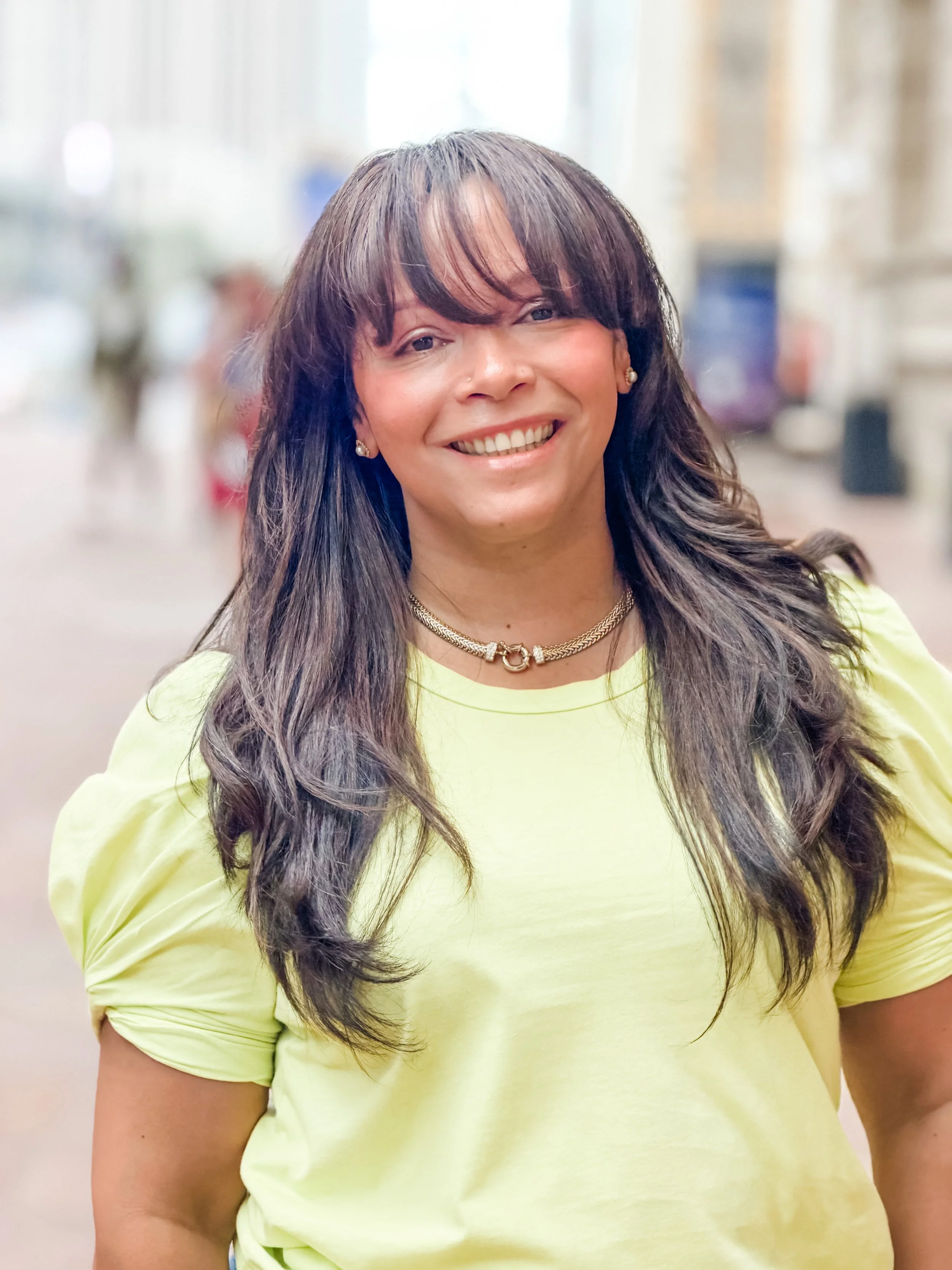 Bianca Reyes is smiling against a blurry, street background, with black hair and bangs and a green-yellow blouse.