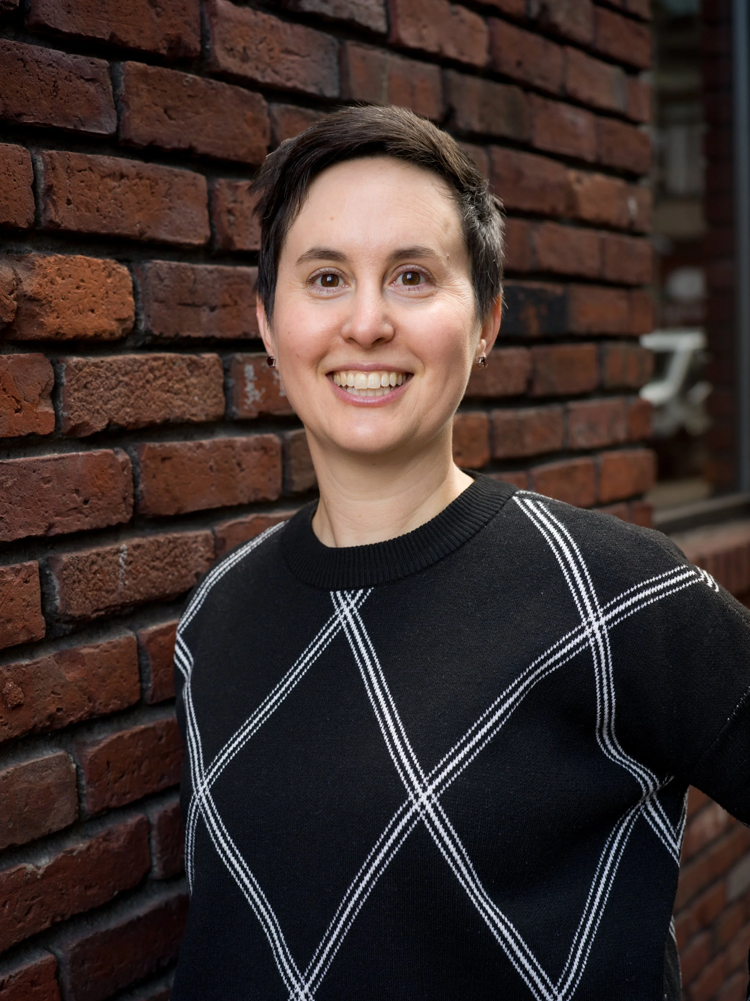 Katrina Magdol is smiling against a brown brick background, wearing a black shirt with a geometric white pattern.