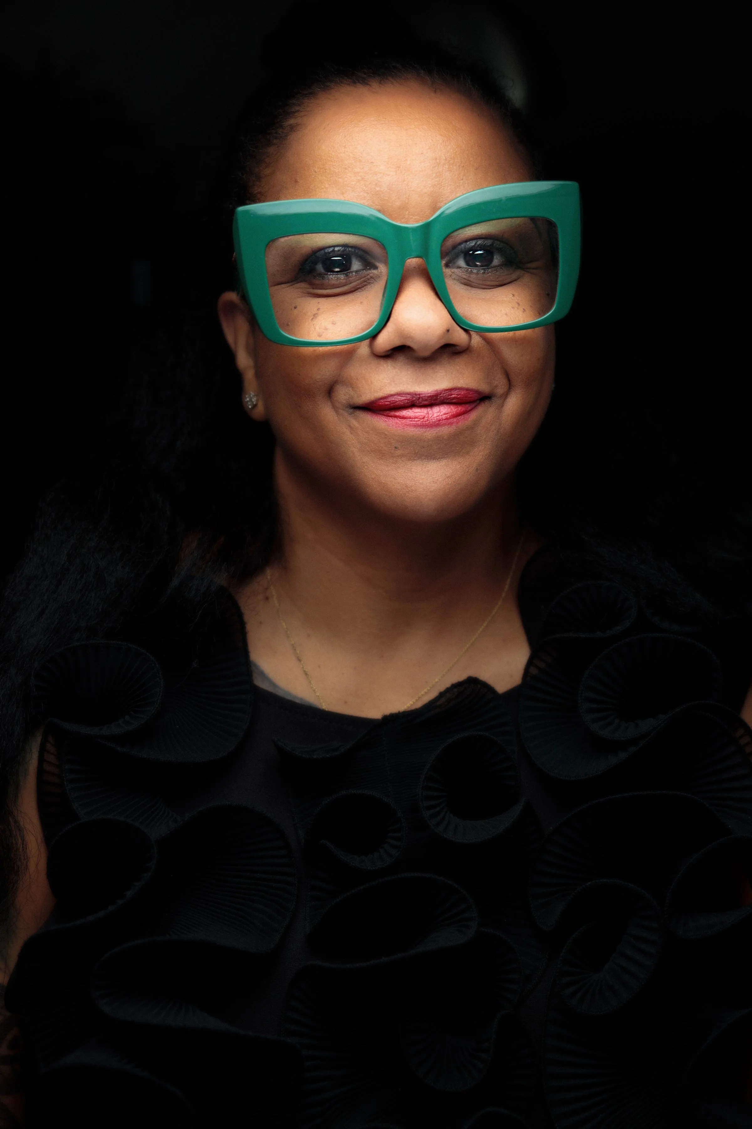 T. Tara Turk-Haynes is smiling against a black background, wearing green glasses and a black shirt.