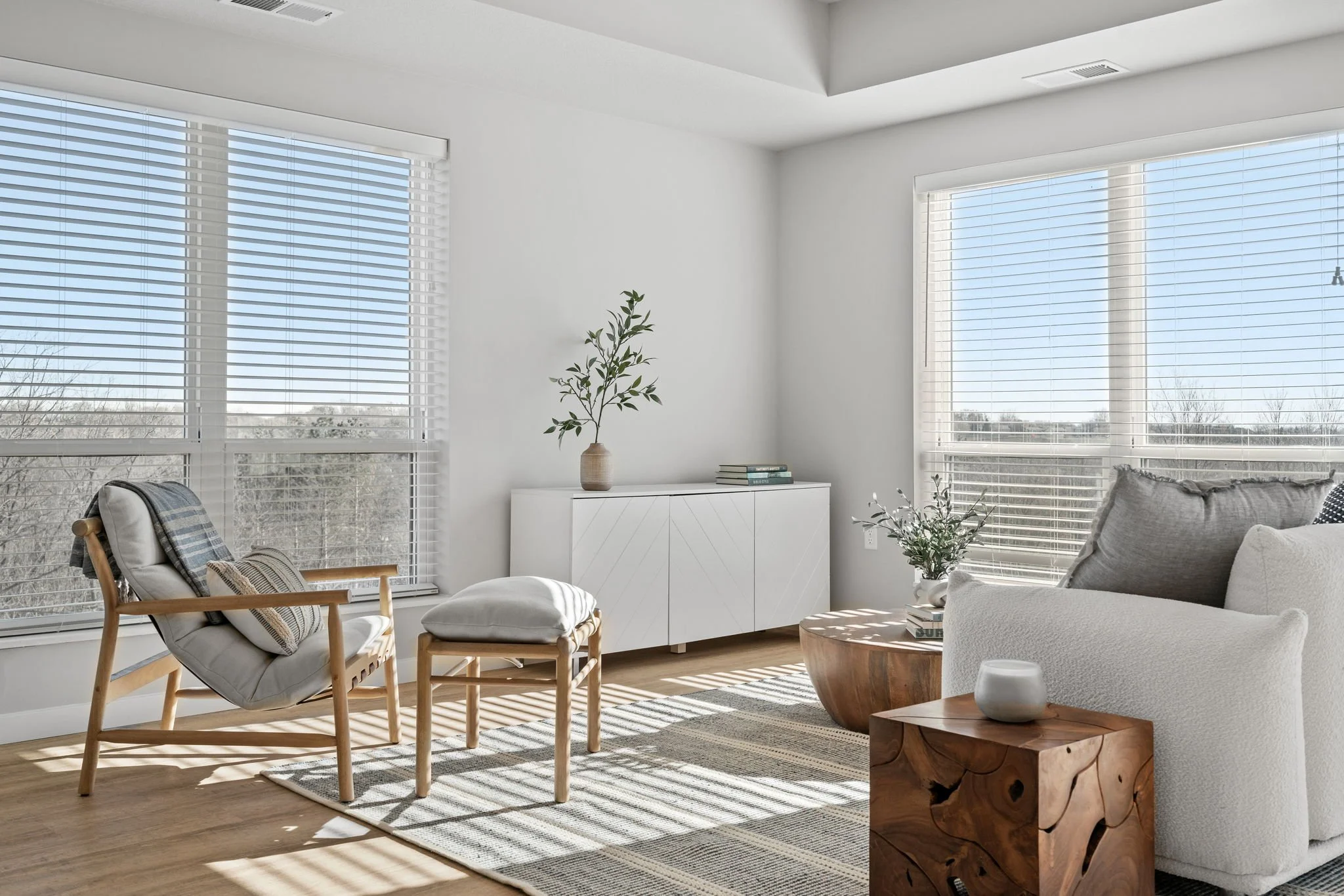 Bright living room with large windows, white walls, wooden furniture, a sofa with cushions, a wooden side table with a white vase, and a white cabinet with plants on top.