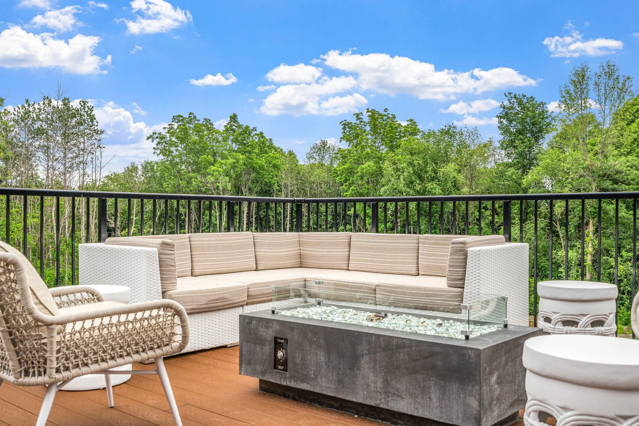 Outdoor patio with wicker and cushioned chairs, a sectional sofa, and a concrete fire pit table surrounded by a black metal railing overlooking a green forest under a partly cloudy blue sky.